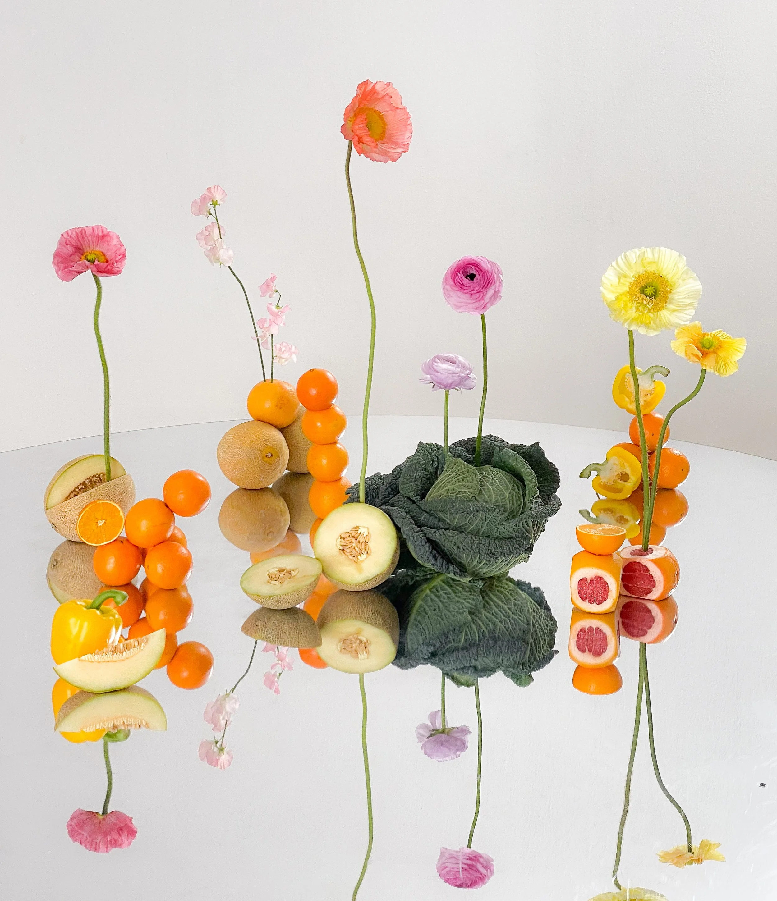 Arrangement of flowers and fruits, including poppy flowers, cantaloupe, oranges, and bell peppers, with reflections on a mirrored surface.
