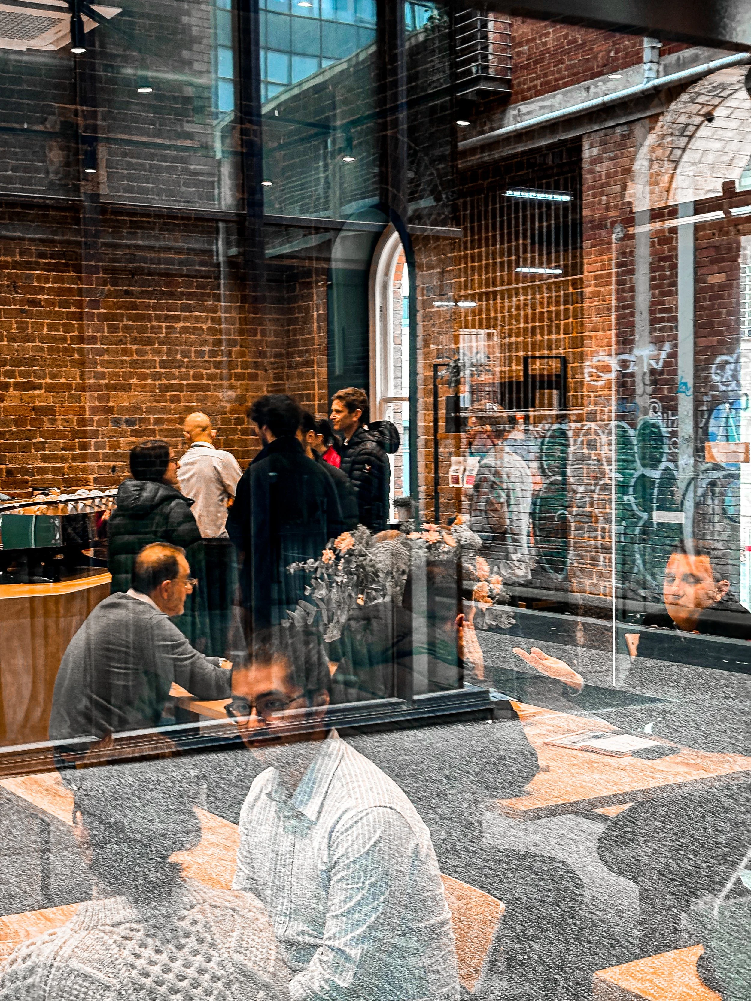 Inside the bustling cafe, with reflections of people outside on the glass window showing some graffiti on a brick wall in the cool Collingwood lane