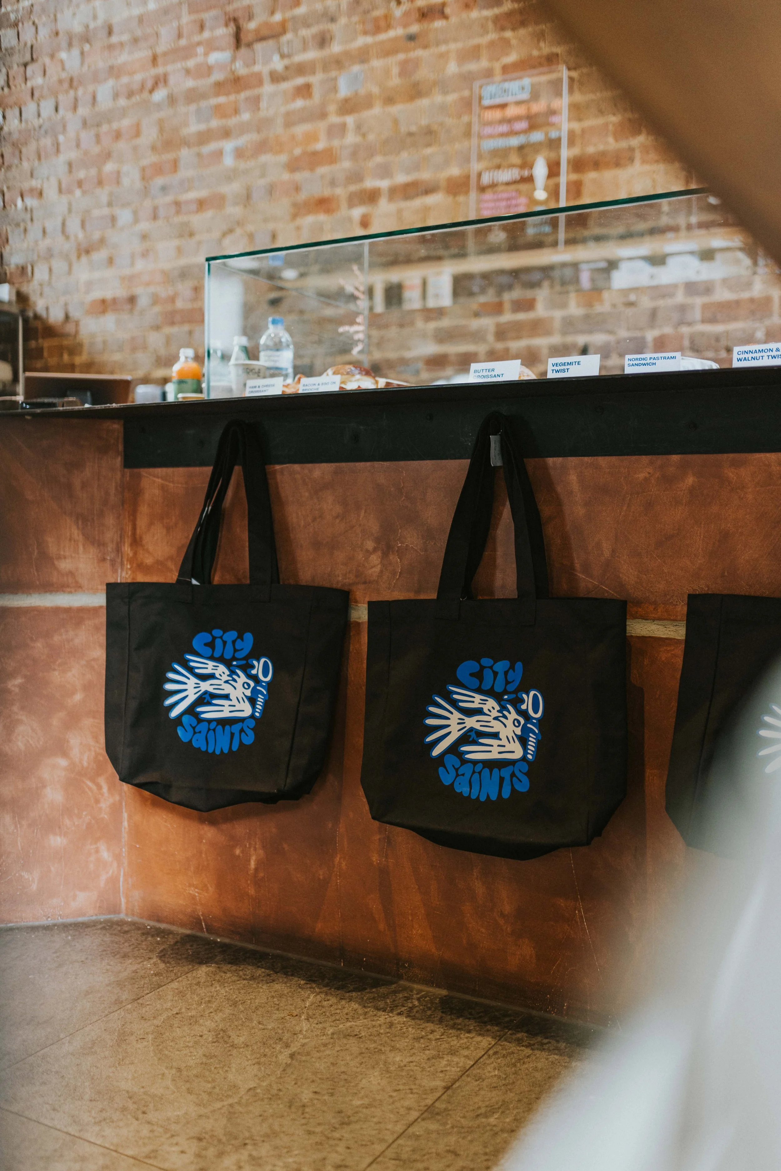 Two black City Saints tote bags hanging on a wooden wall beneath a glass display case