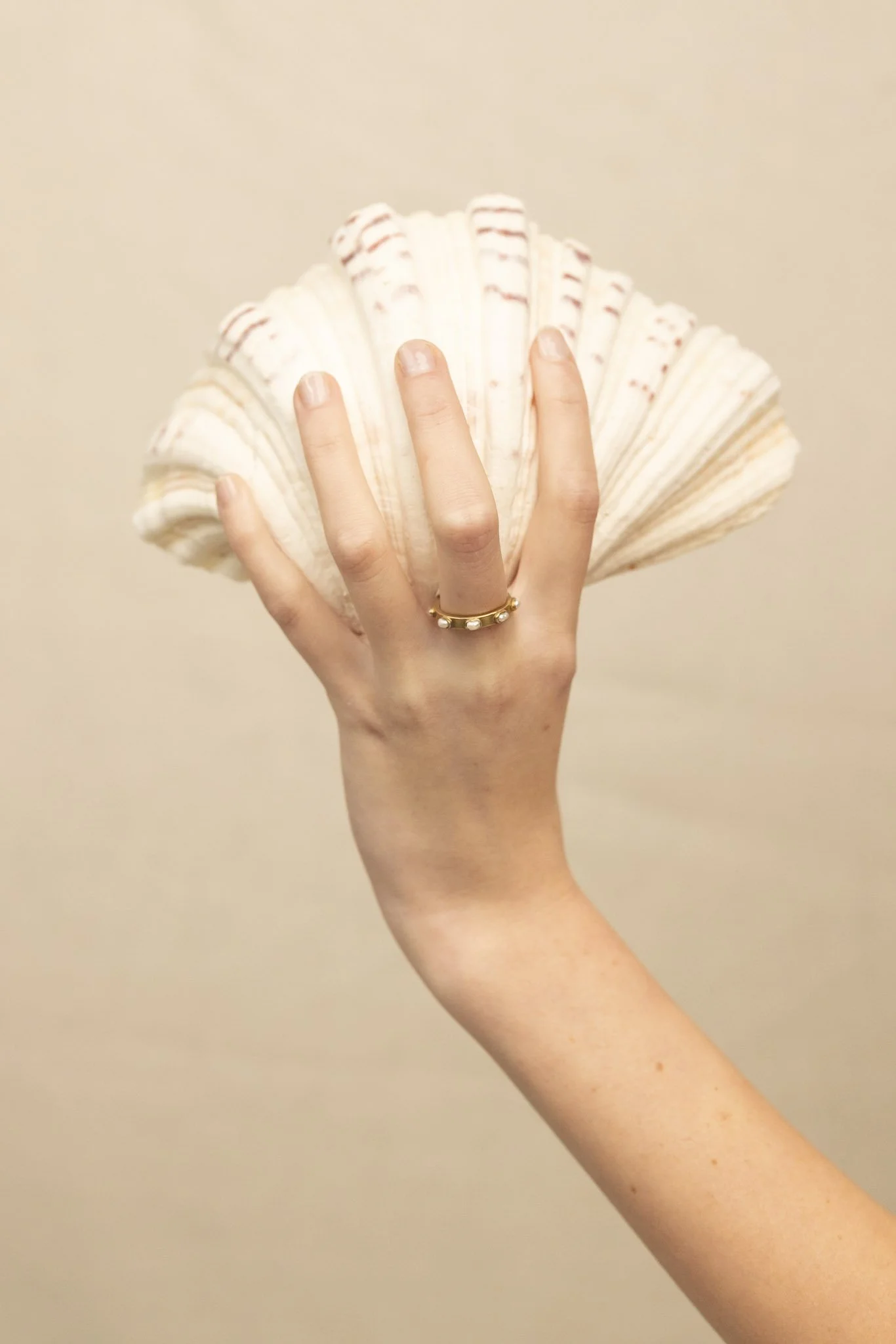 A hand with a gold ring holding a large white seashell against a plain beige background.