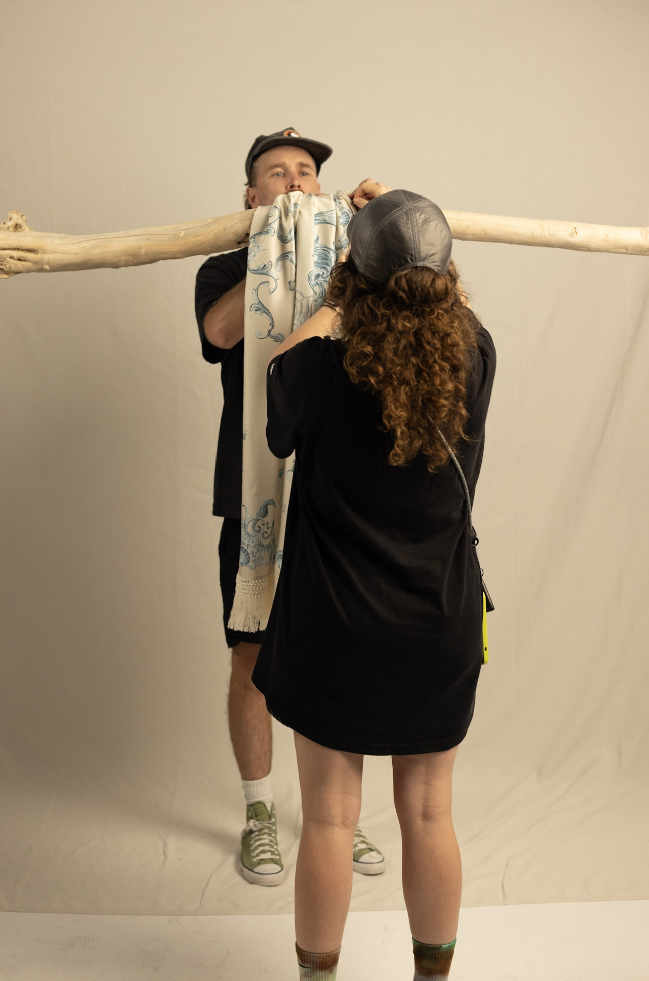 A person with curly hair, wearing a black t-shirt and helmet, is holding a fabric and standing in front of a man with a cap, who is holding a large wooden pole balanced horizontally, with a scarf draped over it. The scene appears to be a photo shoot or a performance setup.