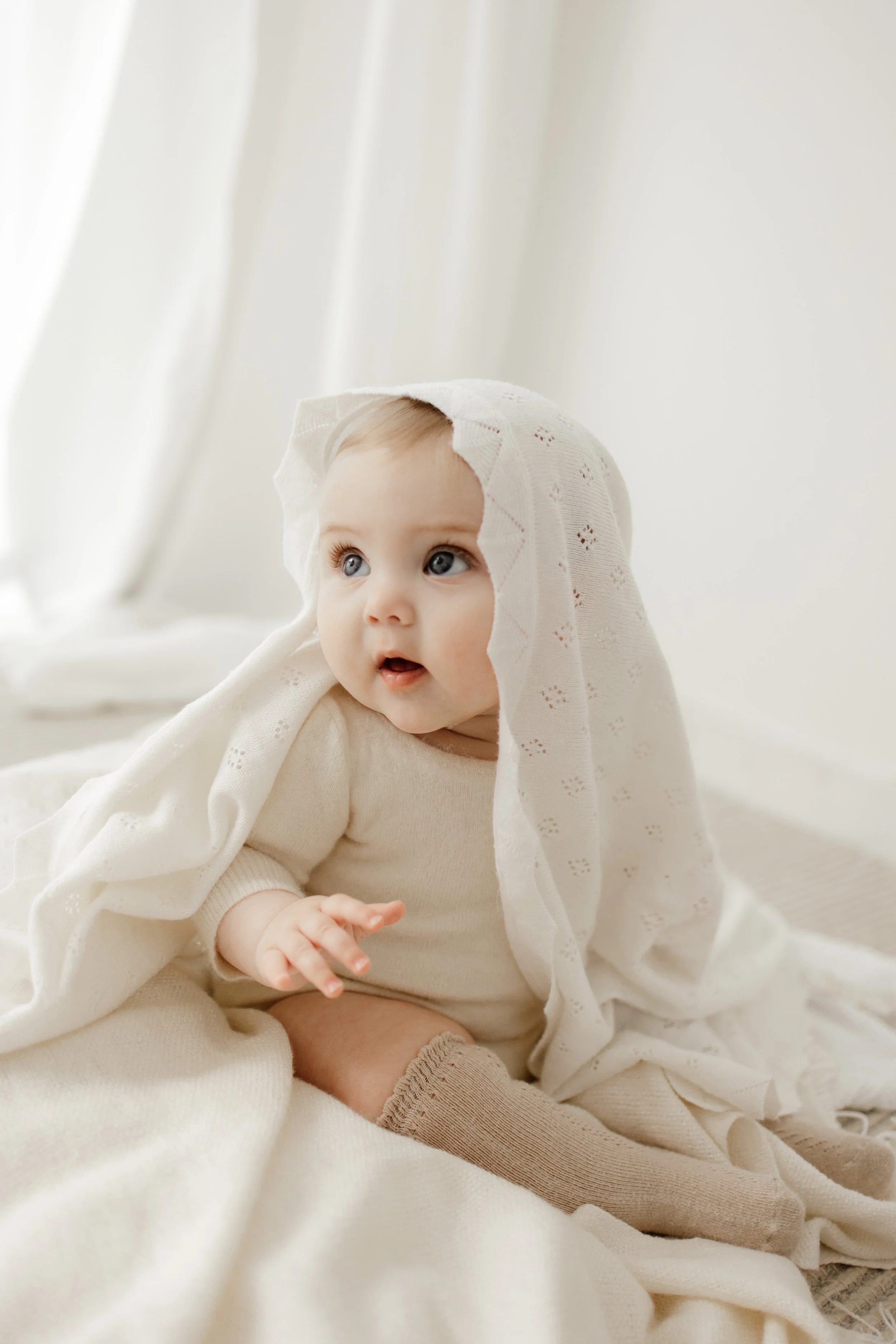 A baby with blue eyes and light hair, wearing a cream-colored outfit and brown socks, sitting on a white blanket with a lace hooded towel draped over their head in a bright, minimal room.