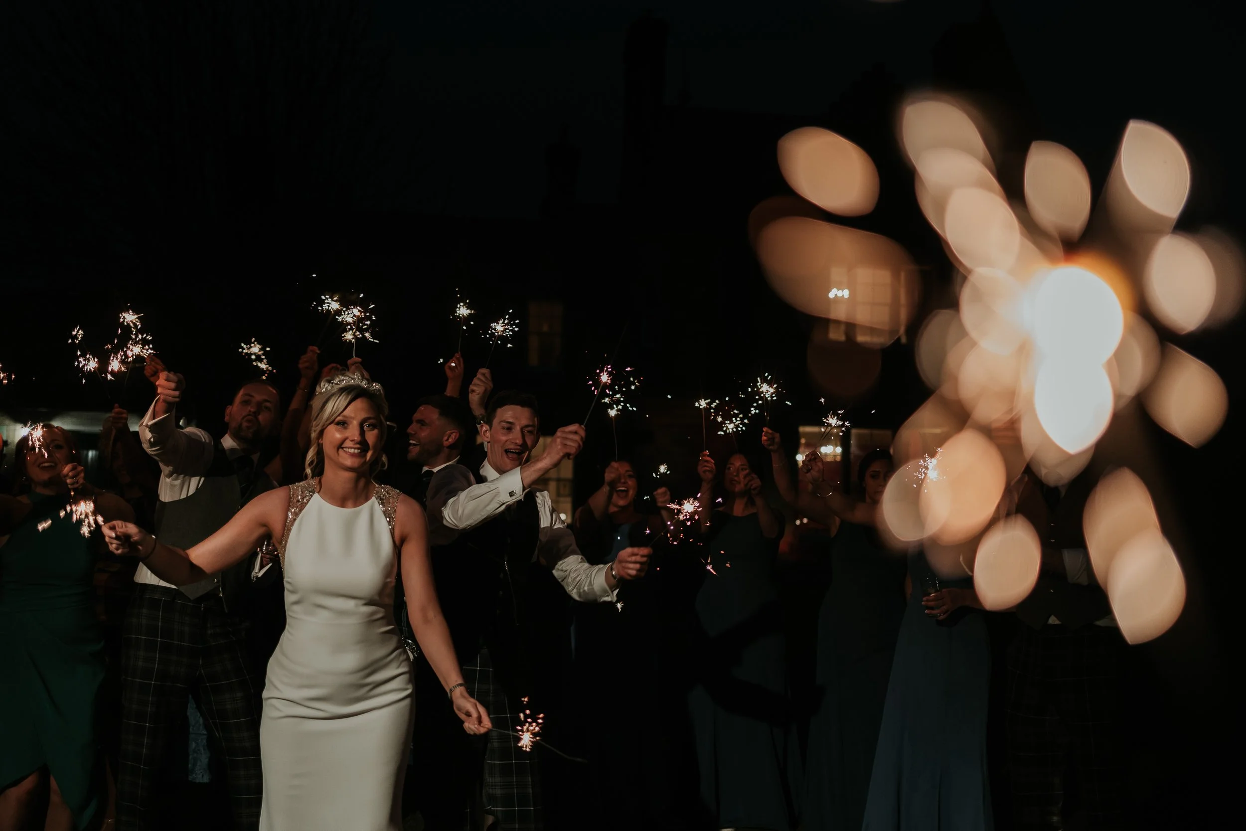 People celebrating at night holding sparklers, some smiling and laughing, with a bokeh effect of lights in the background.