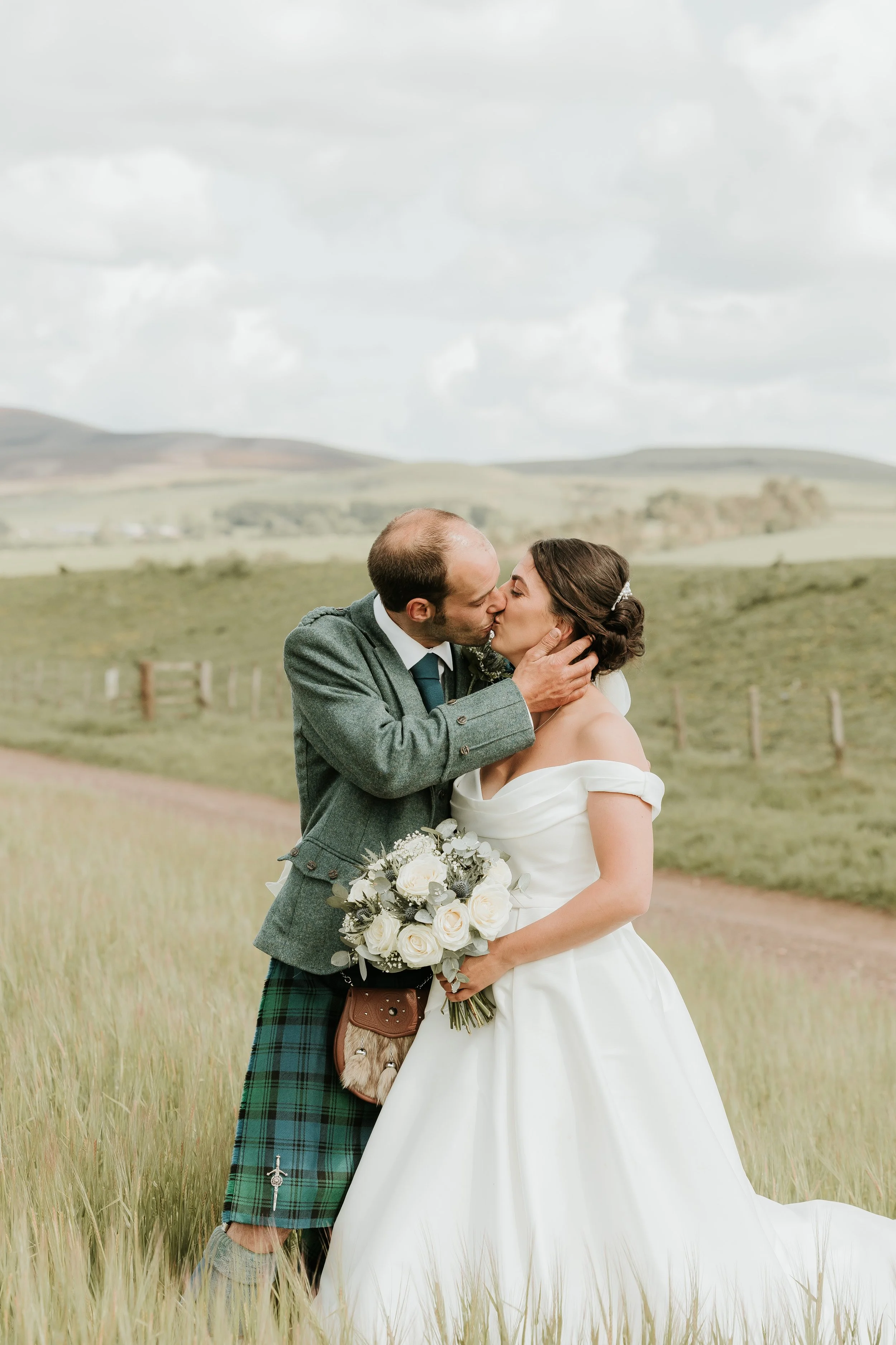 A bride and groom kiss in a grassy field, with the groom wearing a kilt and the bride holding a bouquet of white roses.