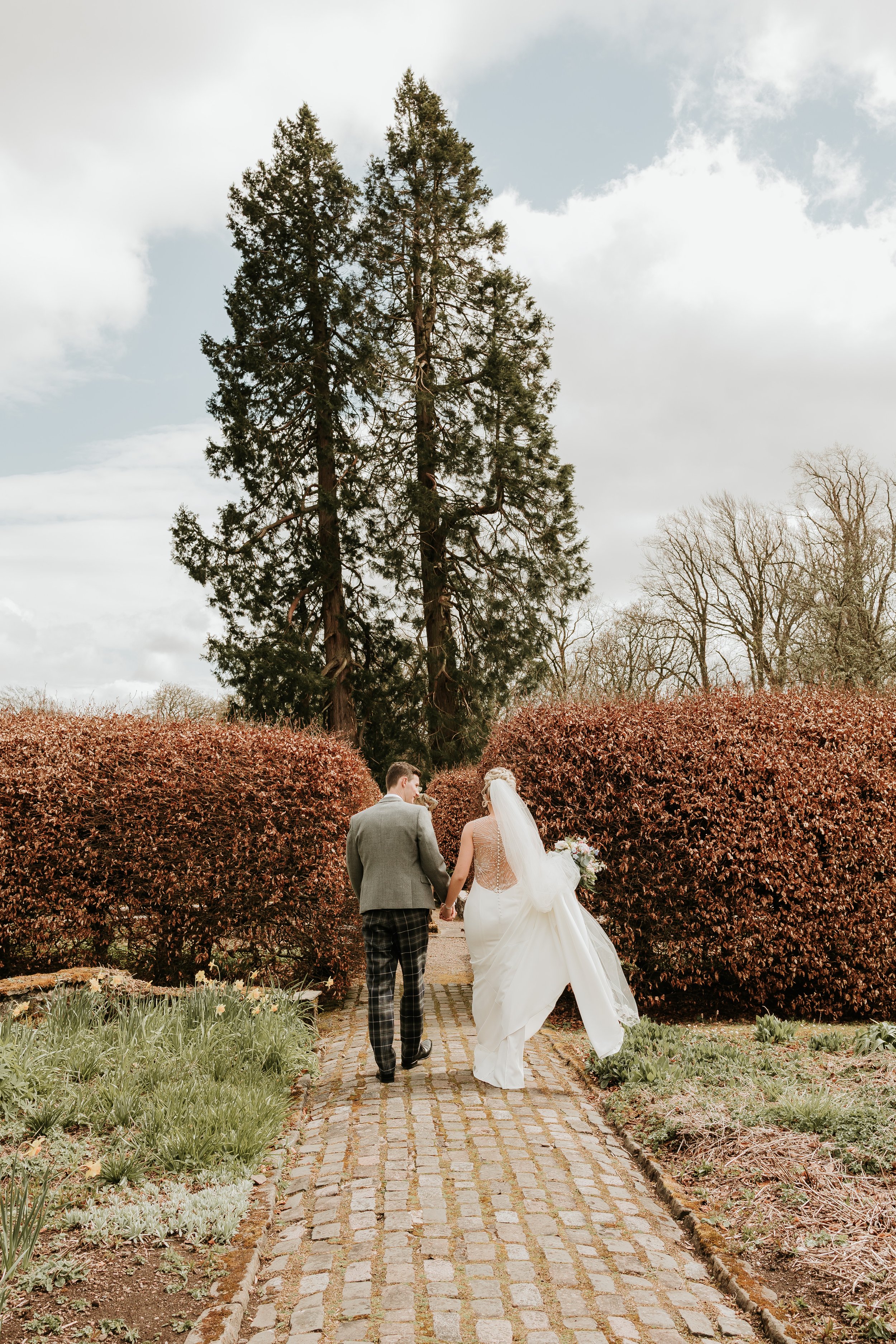 Bride and groom walking hand in hand on a cobblestone path in a garden, surrounded by red bushes and tall trees under a partly cloudy sky.