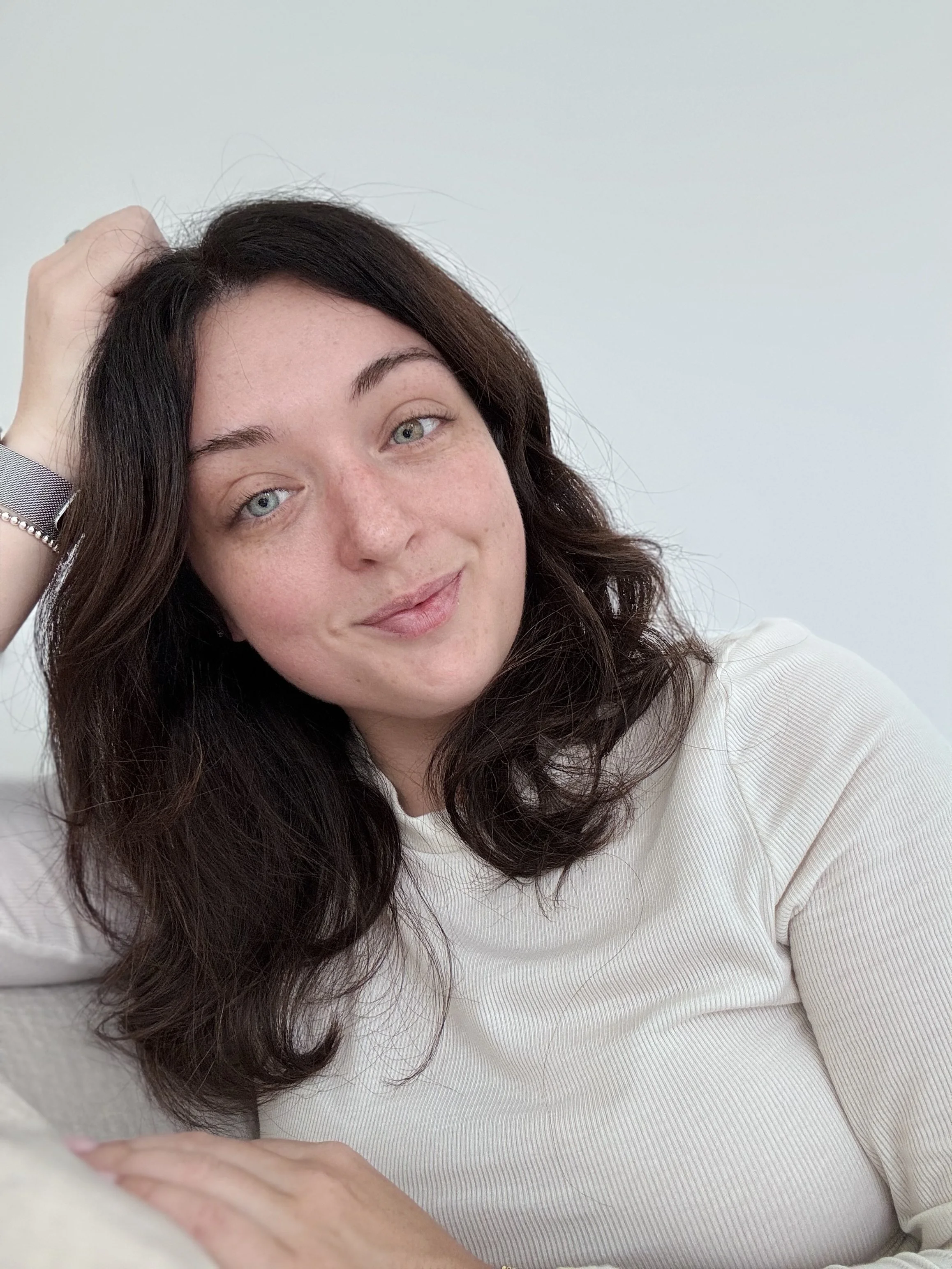 A young woman with long dark hair and light complexion smiling, resting her head on her left hand, wearing a light-colored top, with a plain light background.