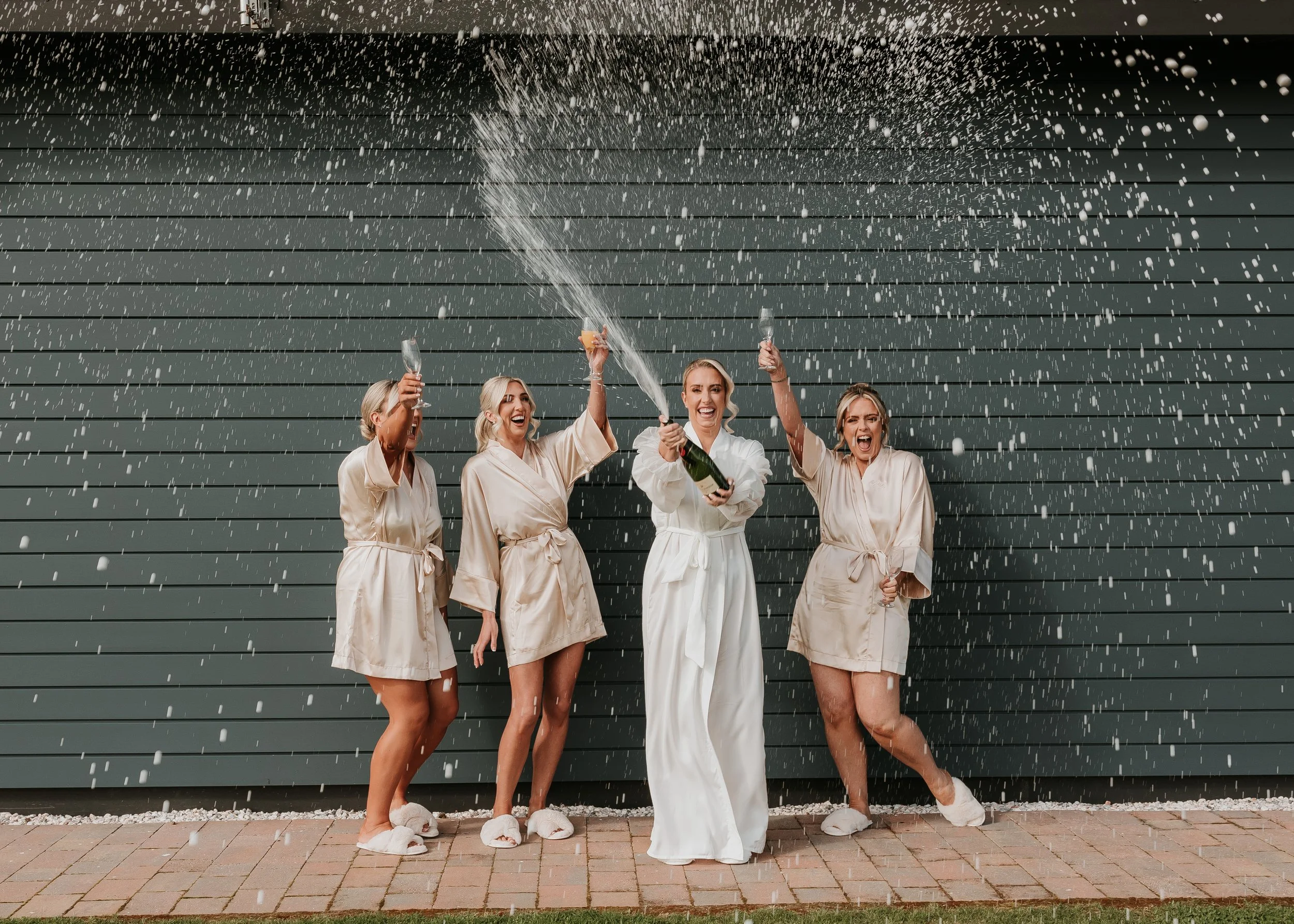 Group of four women in cream-colored robes celebrating with champagne, spraying champagne in front of dark green horizontal siding wall, on brick patio with grass.
