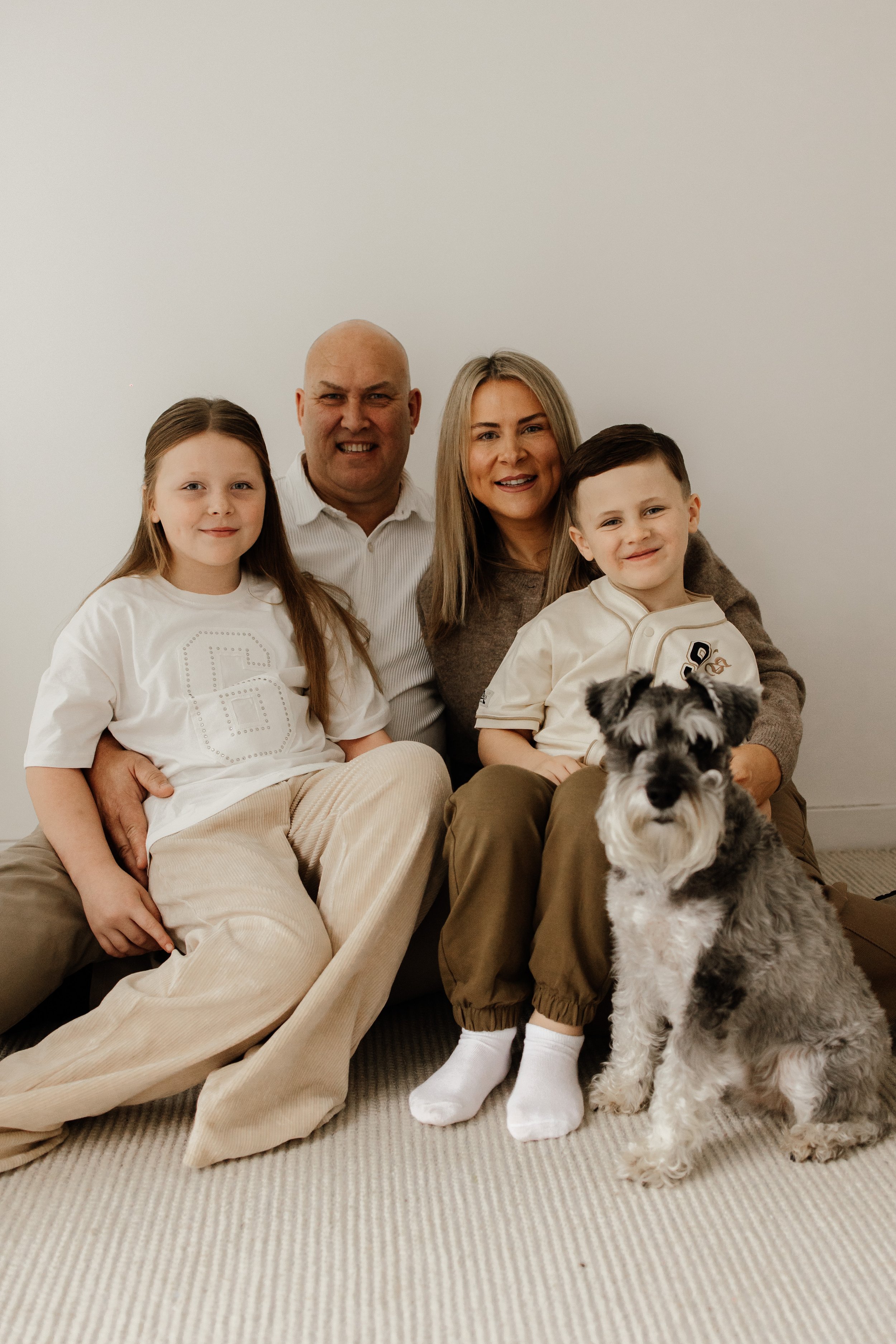 A family of five with a dog sitting together on a beige carpet against a plain white wall. The family includes a man, woman, two children, and a Schnauzer dog.