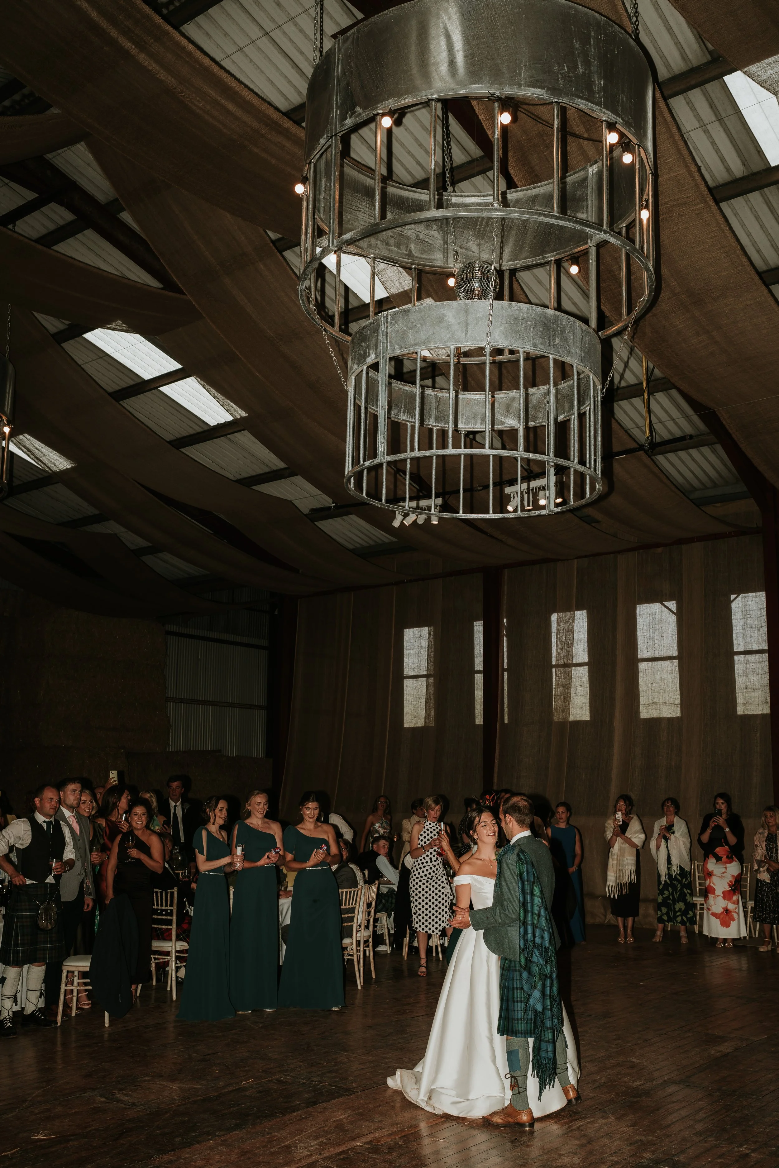 A bride and groom dance together in the center of a rustic wedding reception hall, surrounded by guests watching and taking photos.