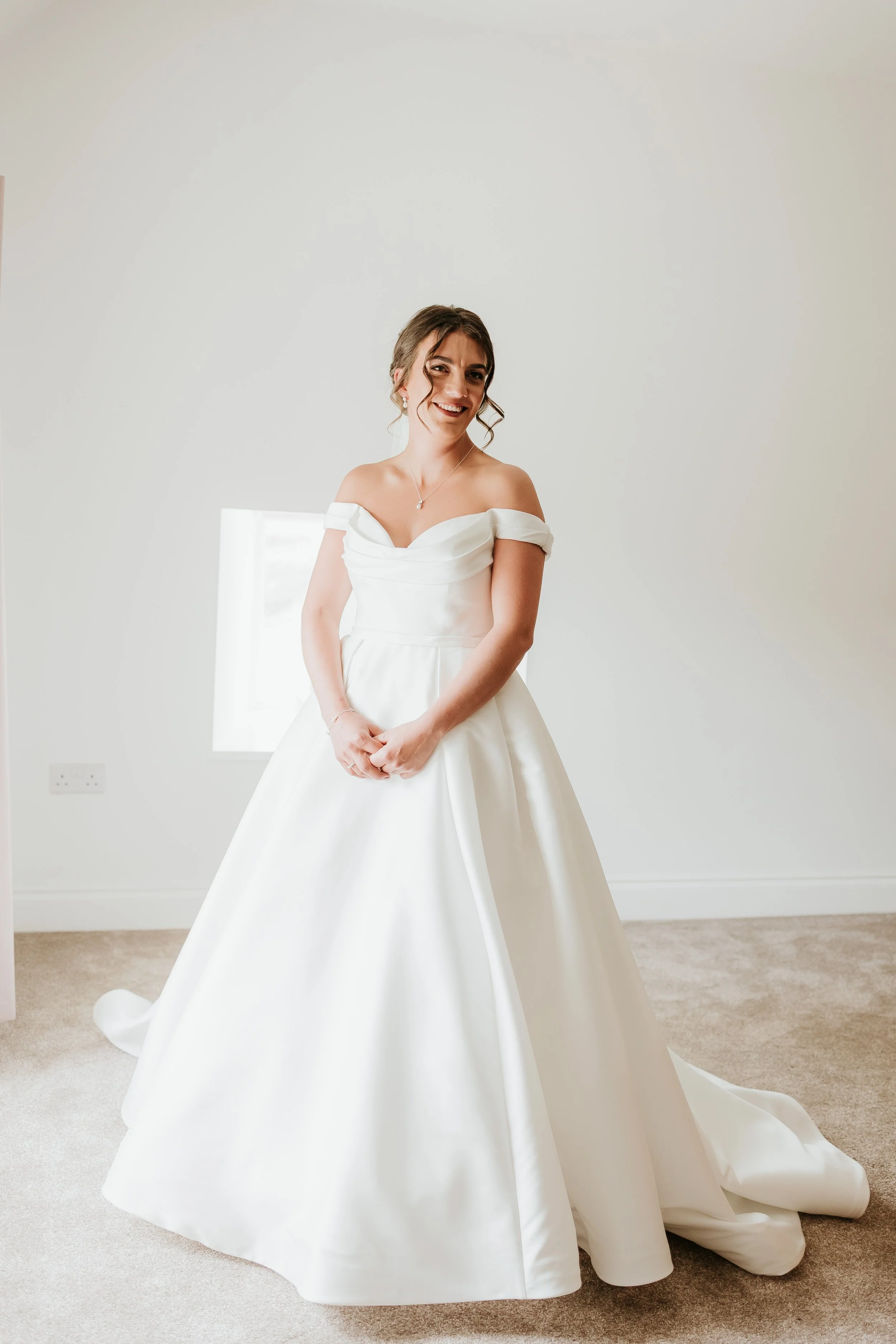 A woman in a white wedding dress standing in a minimal white room, smiling, with curly hair and a necklace.