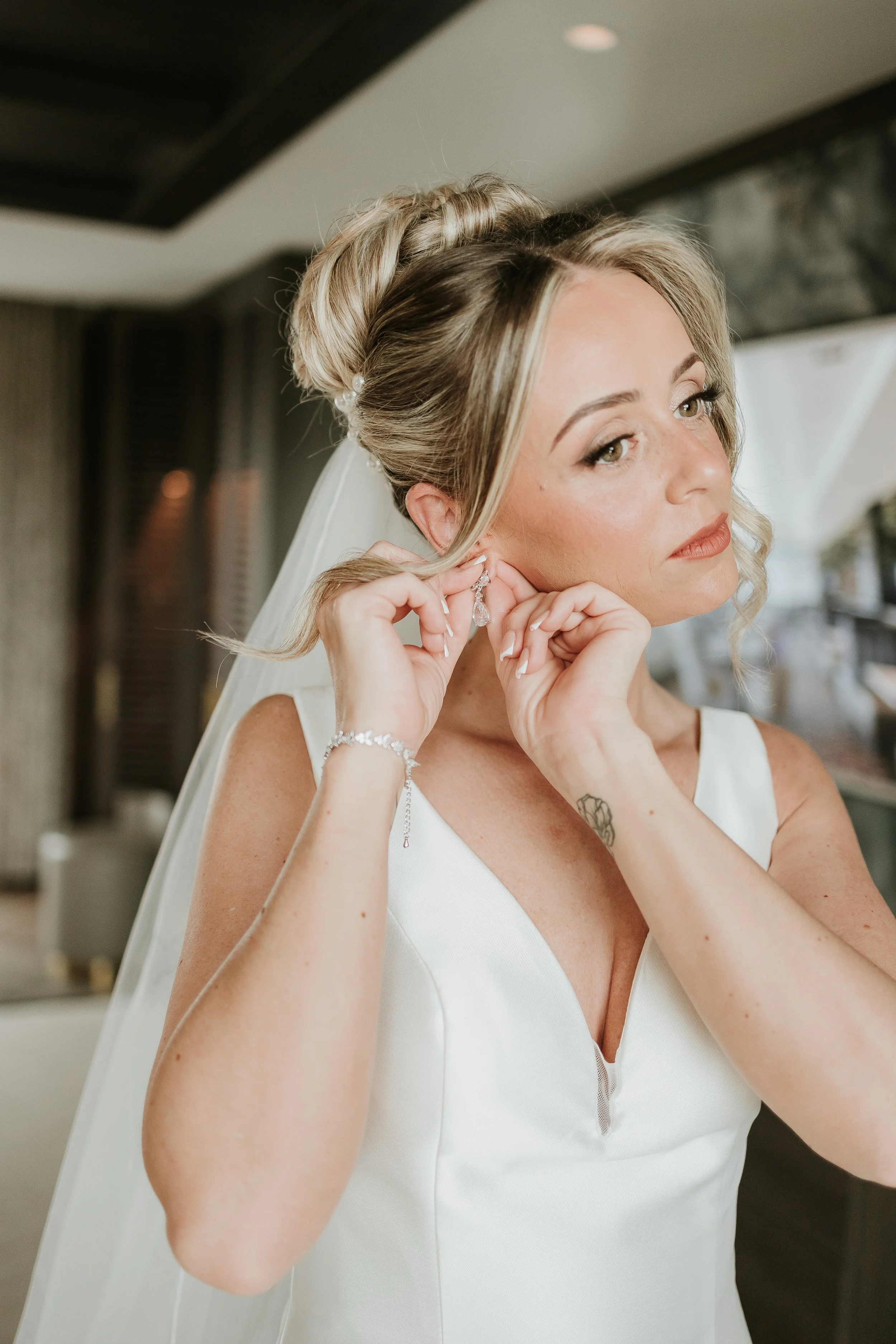 A bride with blonde hair styled in an updo wearing earrings, a bracelet, and a white wedding dress, is adjusting her earring.