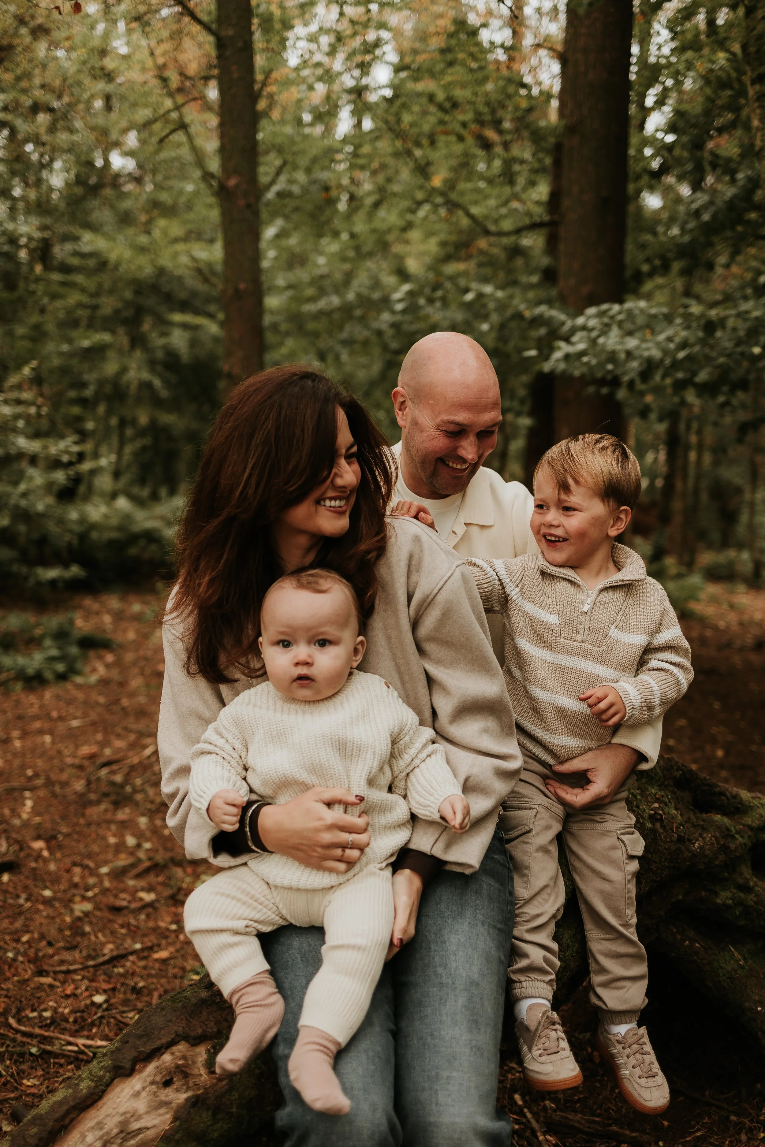 A family of four sitting on a fallen log in a wooded forest, smiling and enjoying a moment together.