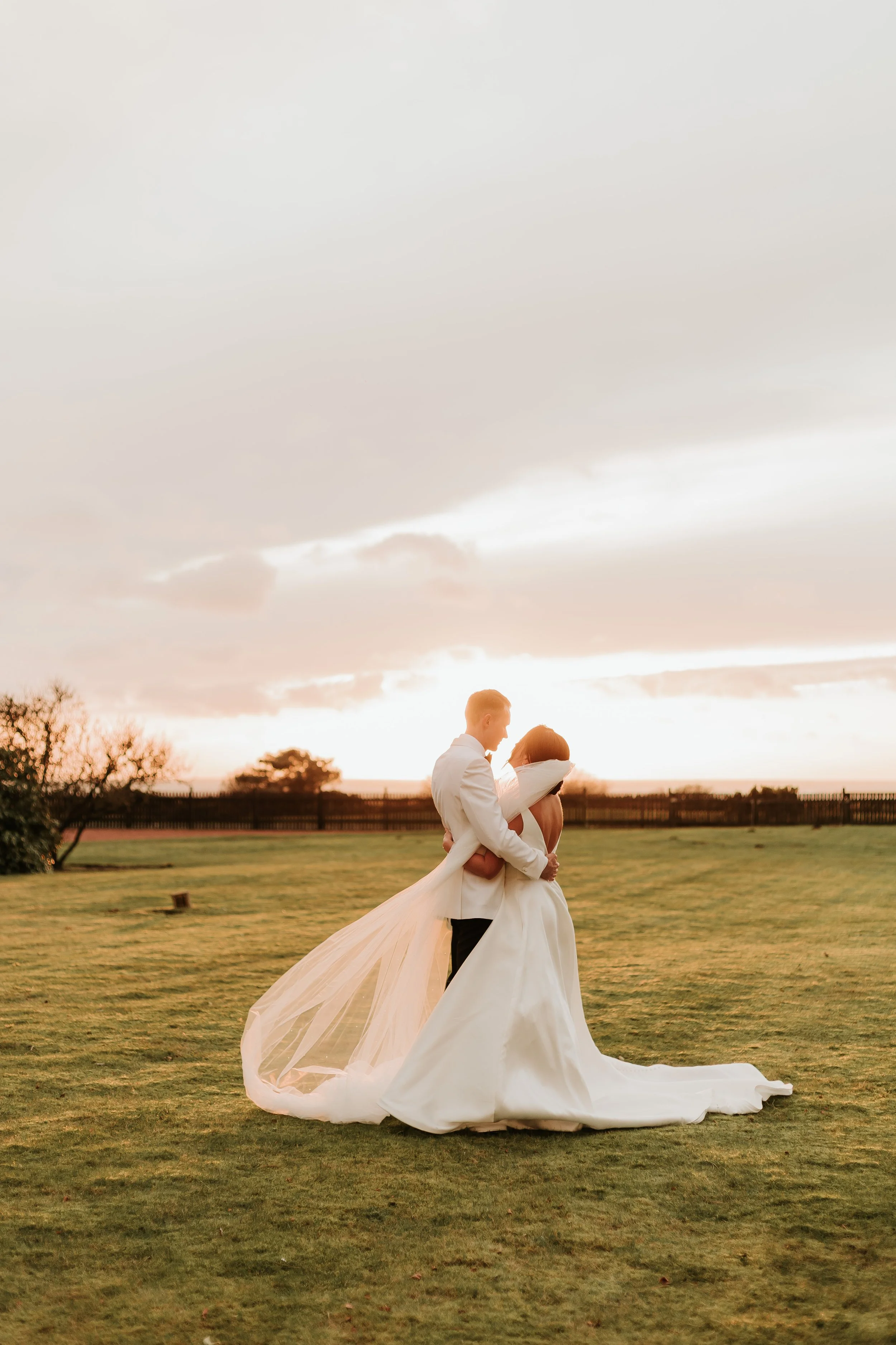 A bride and groom embracing outdoors during sunset, with a sunset sky and grassy field in the background.