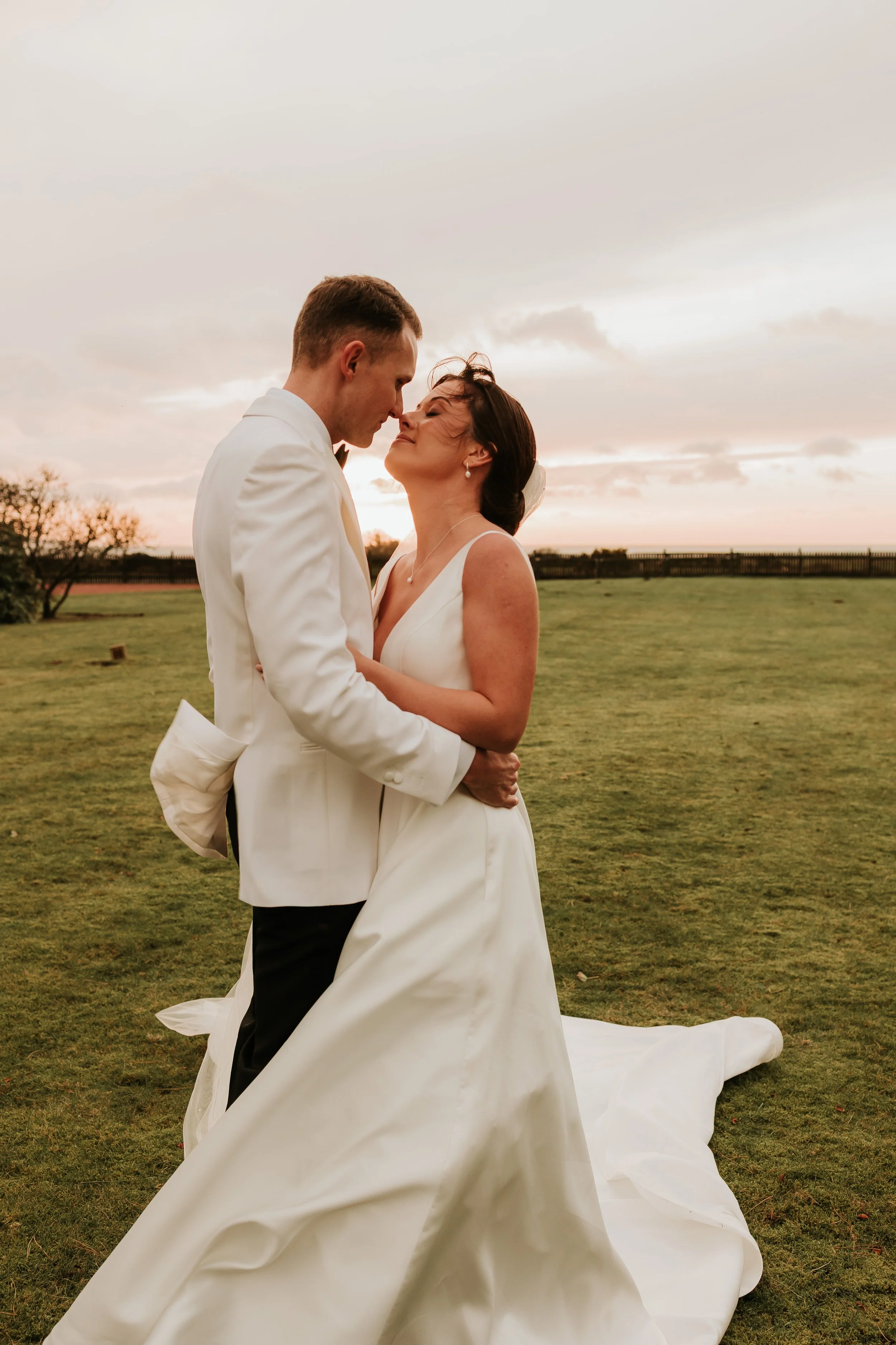 A bride and groom embrace during sunset outdoors, both in white wedding attire, with a grassy field and cloudy sky in the background.