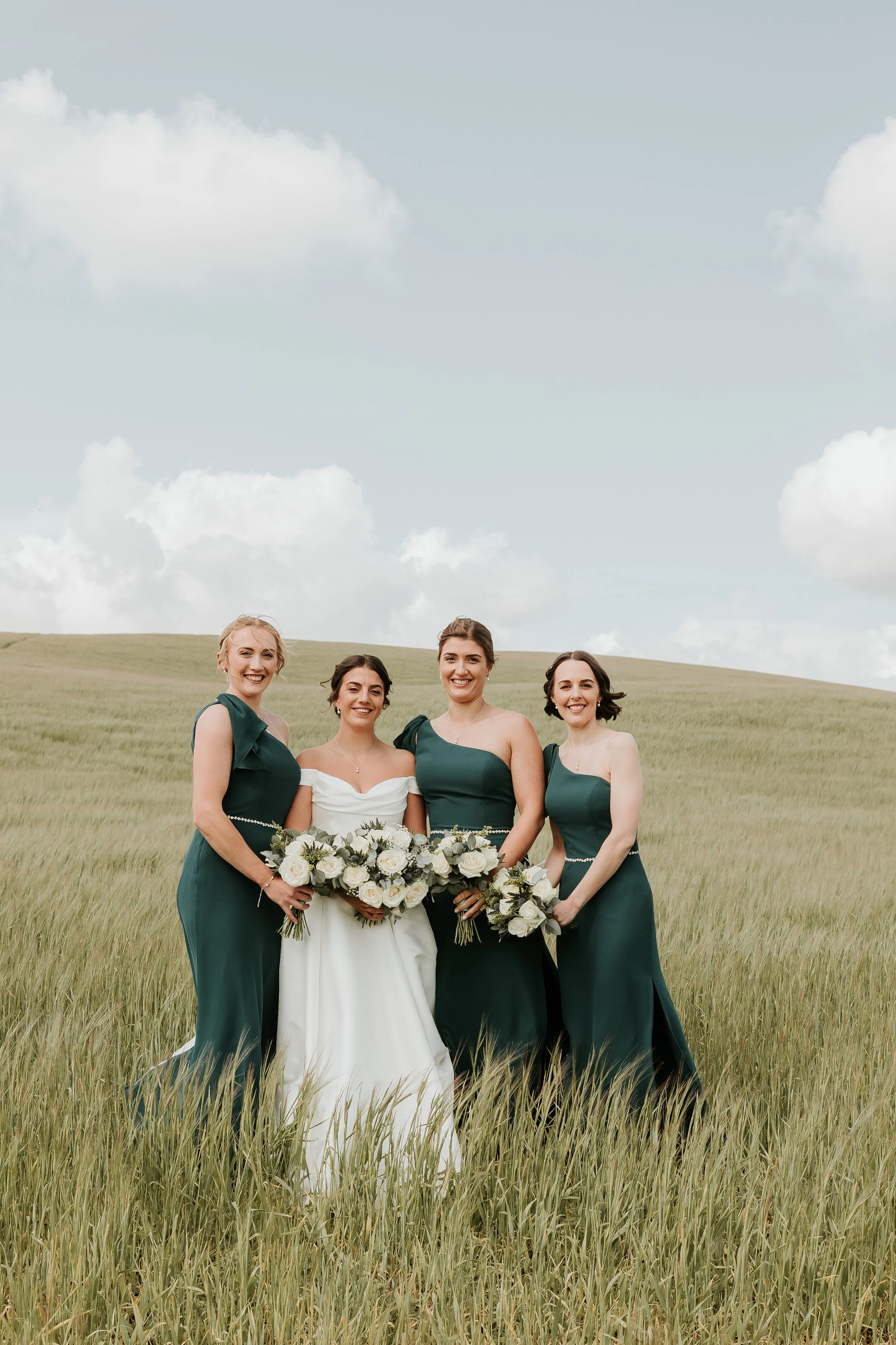 Four women in formal dresses standing in a grassy field, holding bouquets, with a cloudy sky in the background.