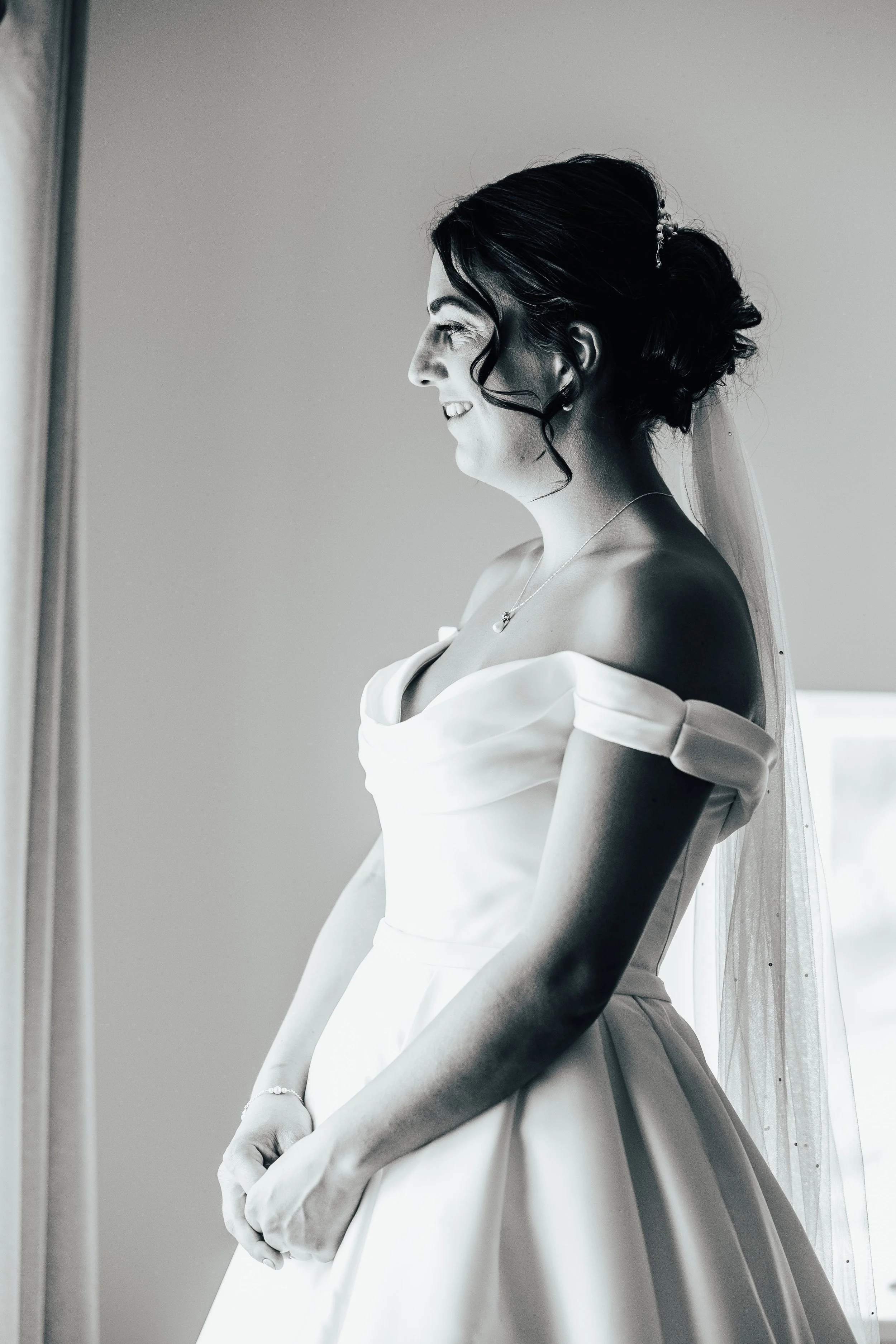 A woman in a wedding dress standing by a window, smiling and looking outside.