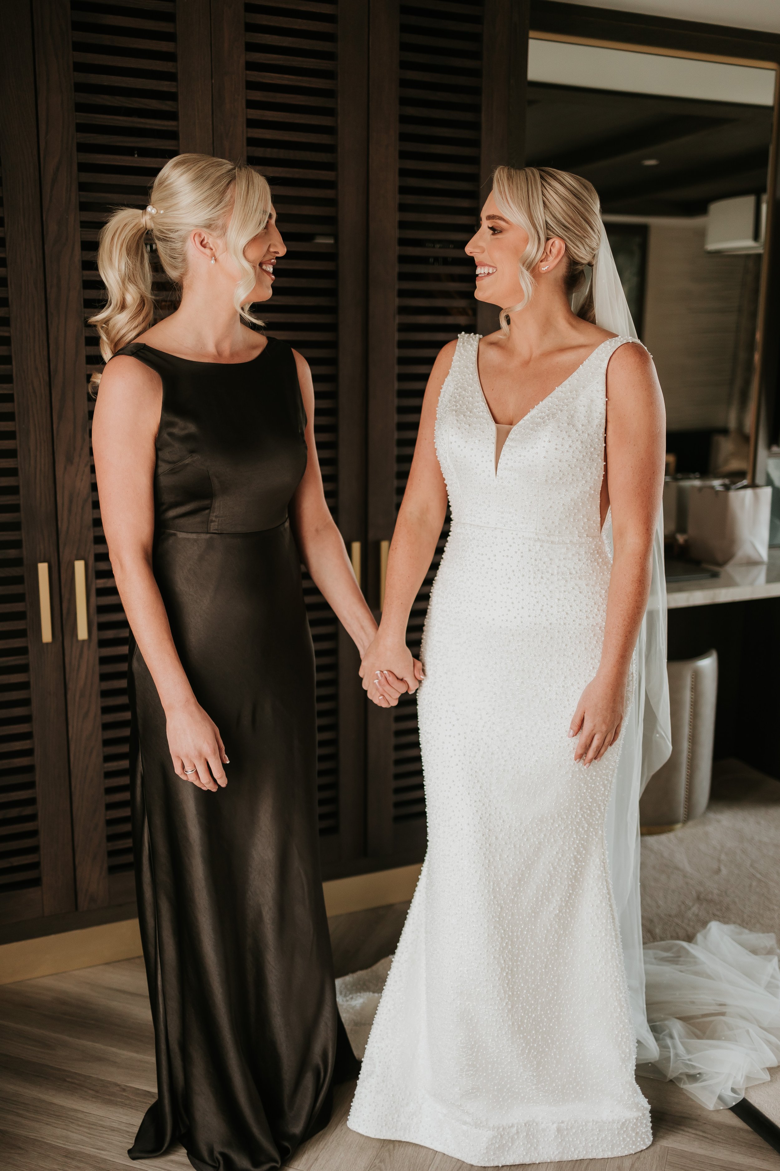 Two women, one in a white wedding dress and the other in a black dress, holding hands and smiling at each other.