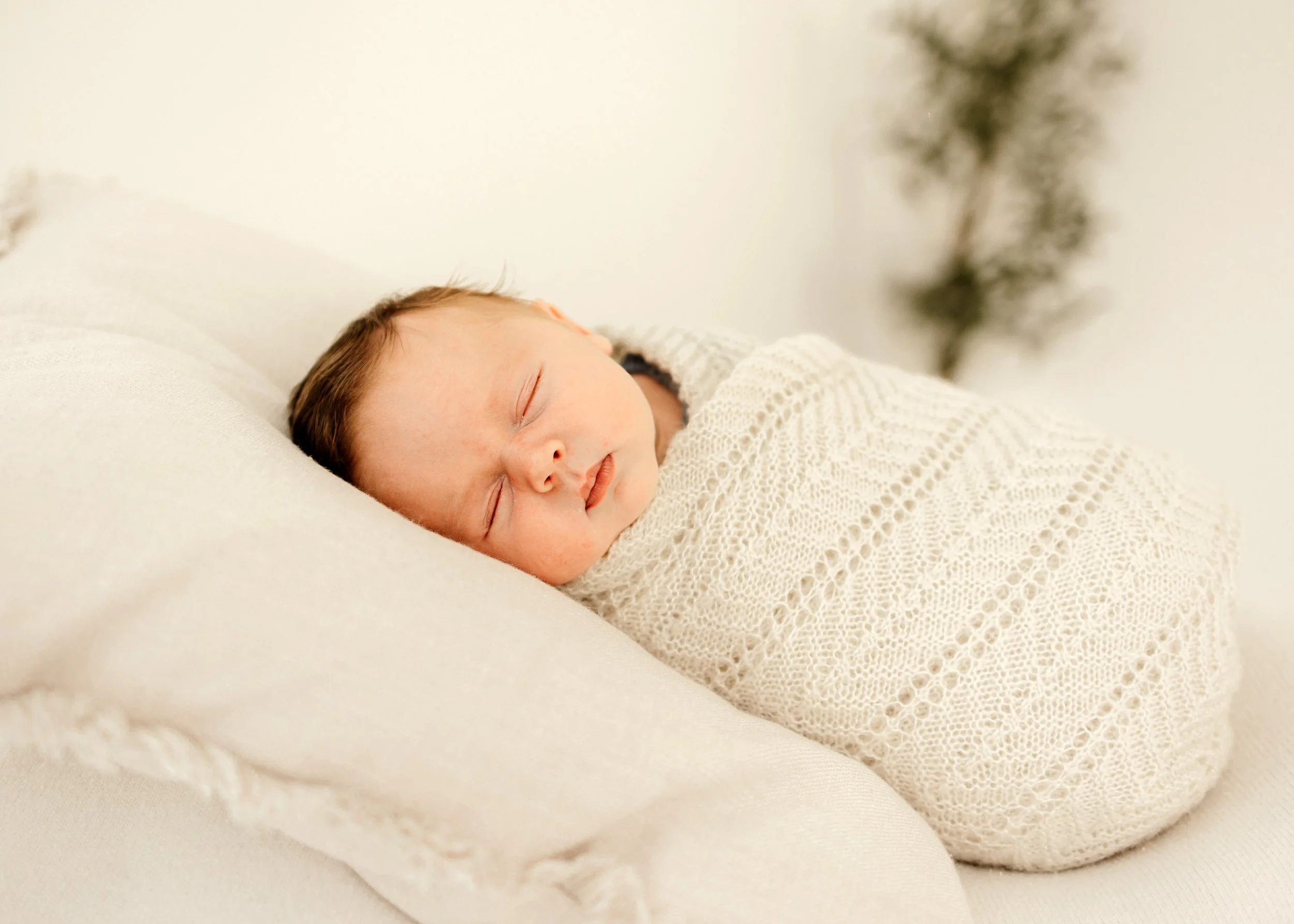 A sleeping baby wrapped in a cream-colored knitted blanket, lying on a cream pillow in a cozy, neutral-toned environment.
