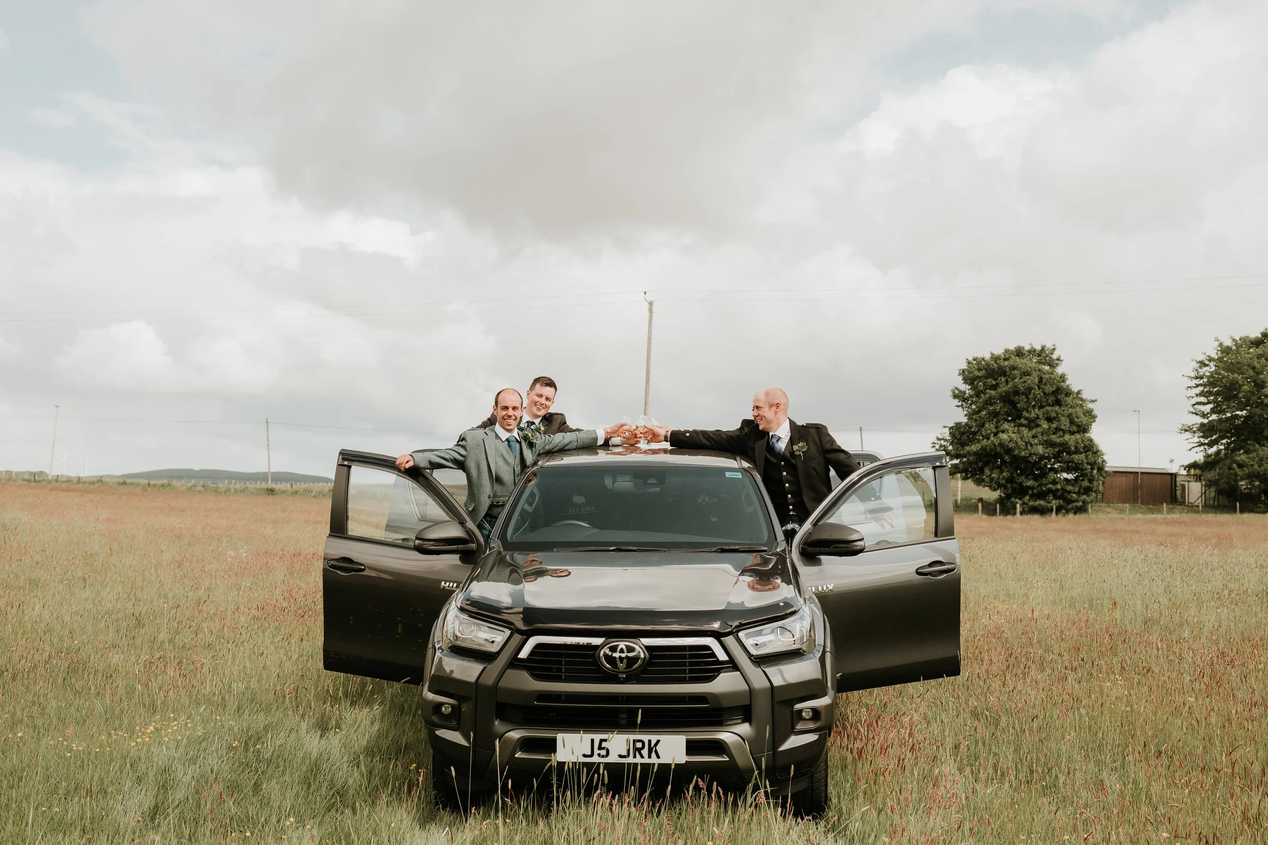 Three men in suits standing through open car doors and on the roof of a black Toyota vehicle in a grassy field, celebrating with drinks and toasting each other, under a cloudy sky.