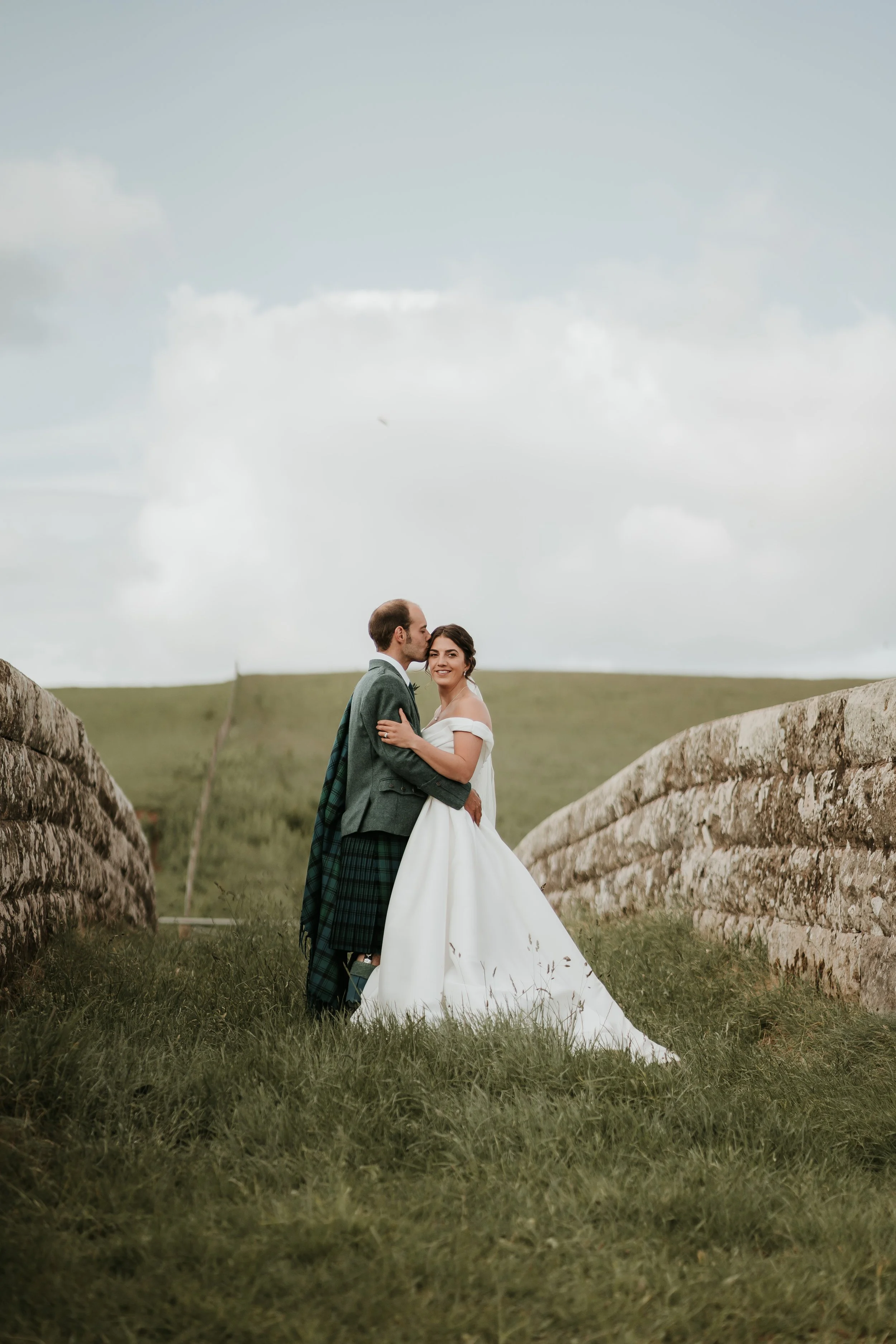 A bride and groom in wedding attire standing between two stone walls on a grassy hill, sharing a kiss and smiling outdoors.
