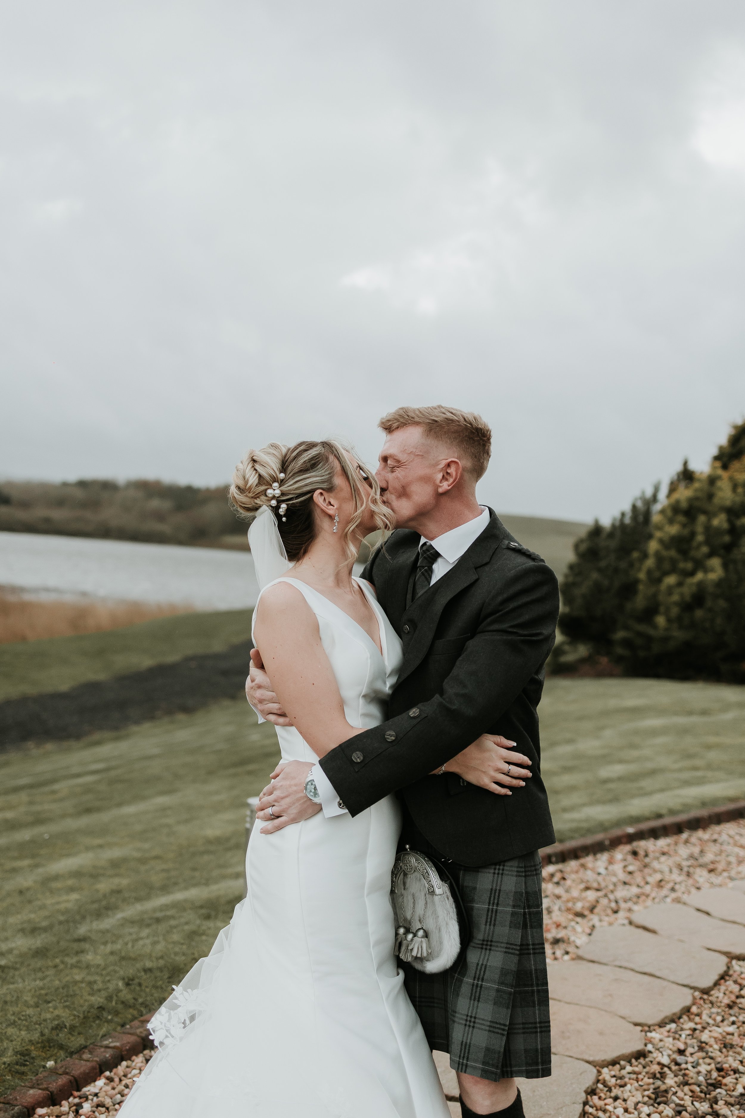 A bride and groom embrace and kiss outdoors on a cloudy day, with a lake, grassy landscape, and trees in the background.