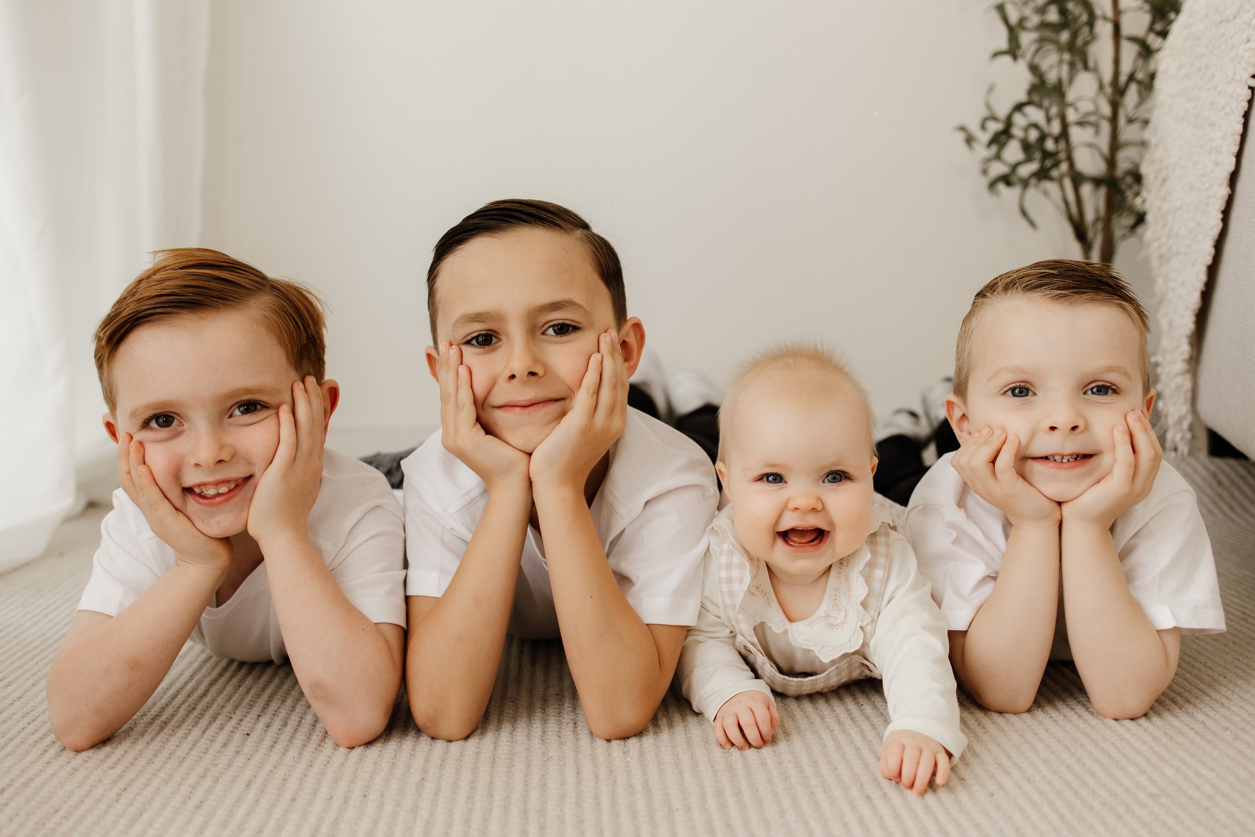 Four children lying on their stomachs on a beige carpet, smiling and resting their faces in their hands, indoors with a potted plant and white curtains in the background.