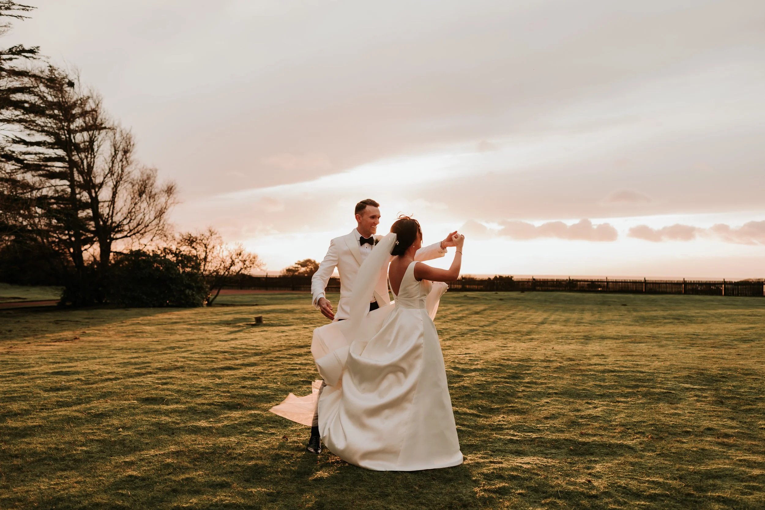 A bride and groom dancing on a grassy field during sunset, the bride in a white gown and the groom in a tuxedo.