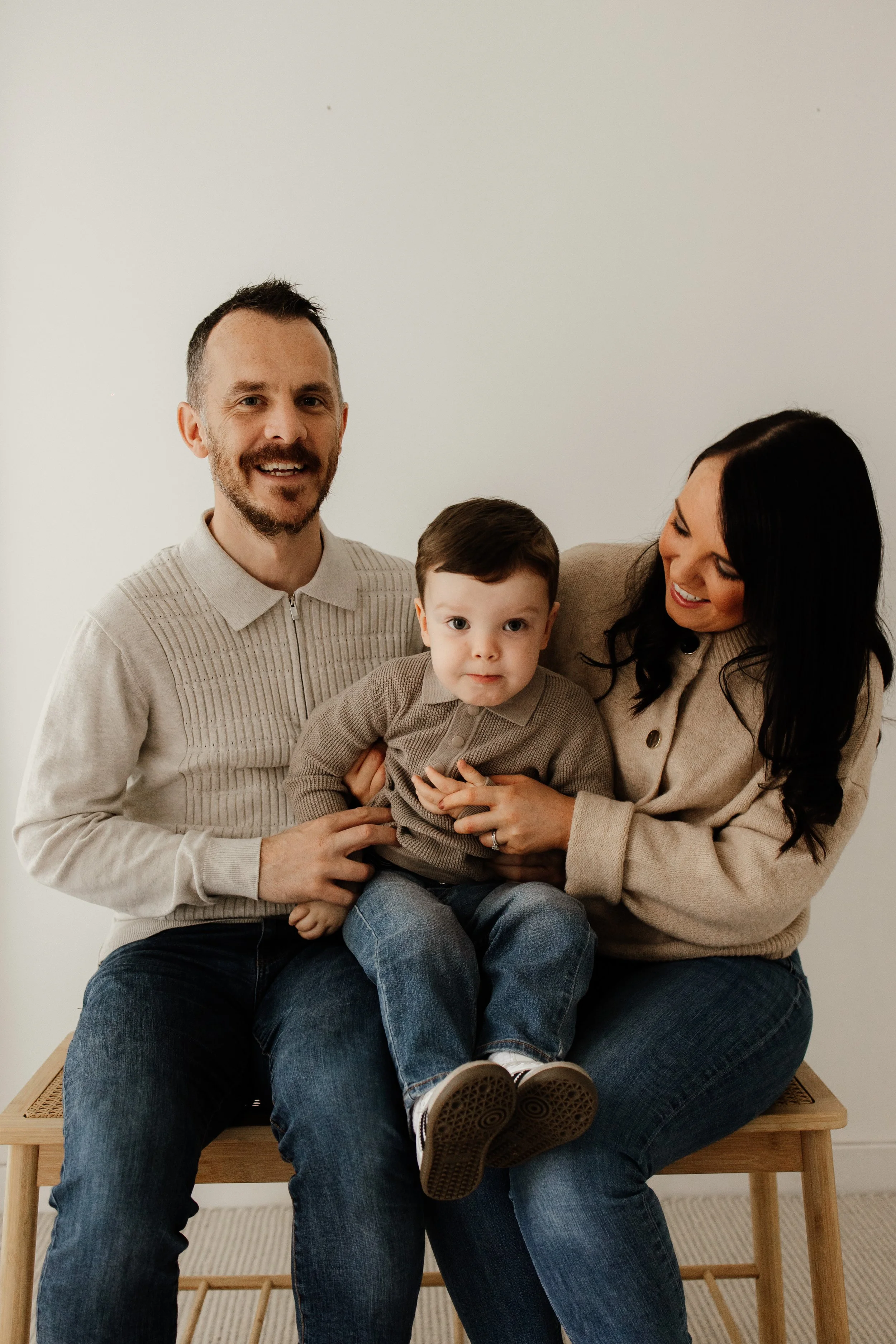A family of three sitting on a wooden bench, smiling and looking at the camera. The man has short dark hair, a beard, and is wearing a light beige sweater. The woman has long dark hair and is wearing a beige jacket. The young boy has short dark hair and is wearing a beige sweater and jeans.