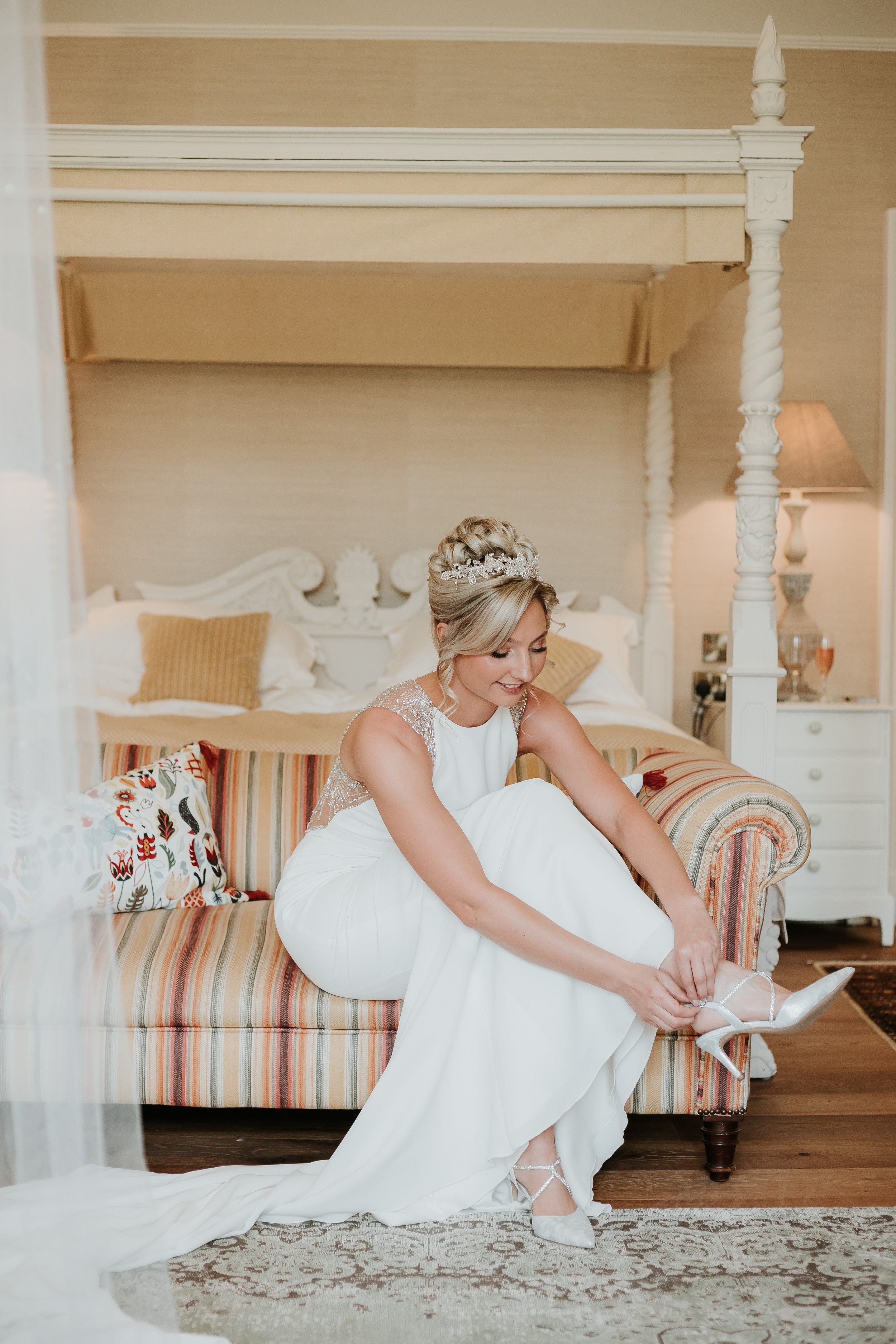 Bride sitting on a striped sofa, putting on her white high heels in a decorated room.