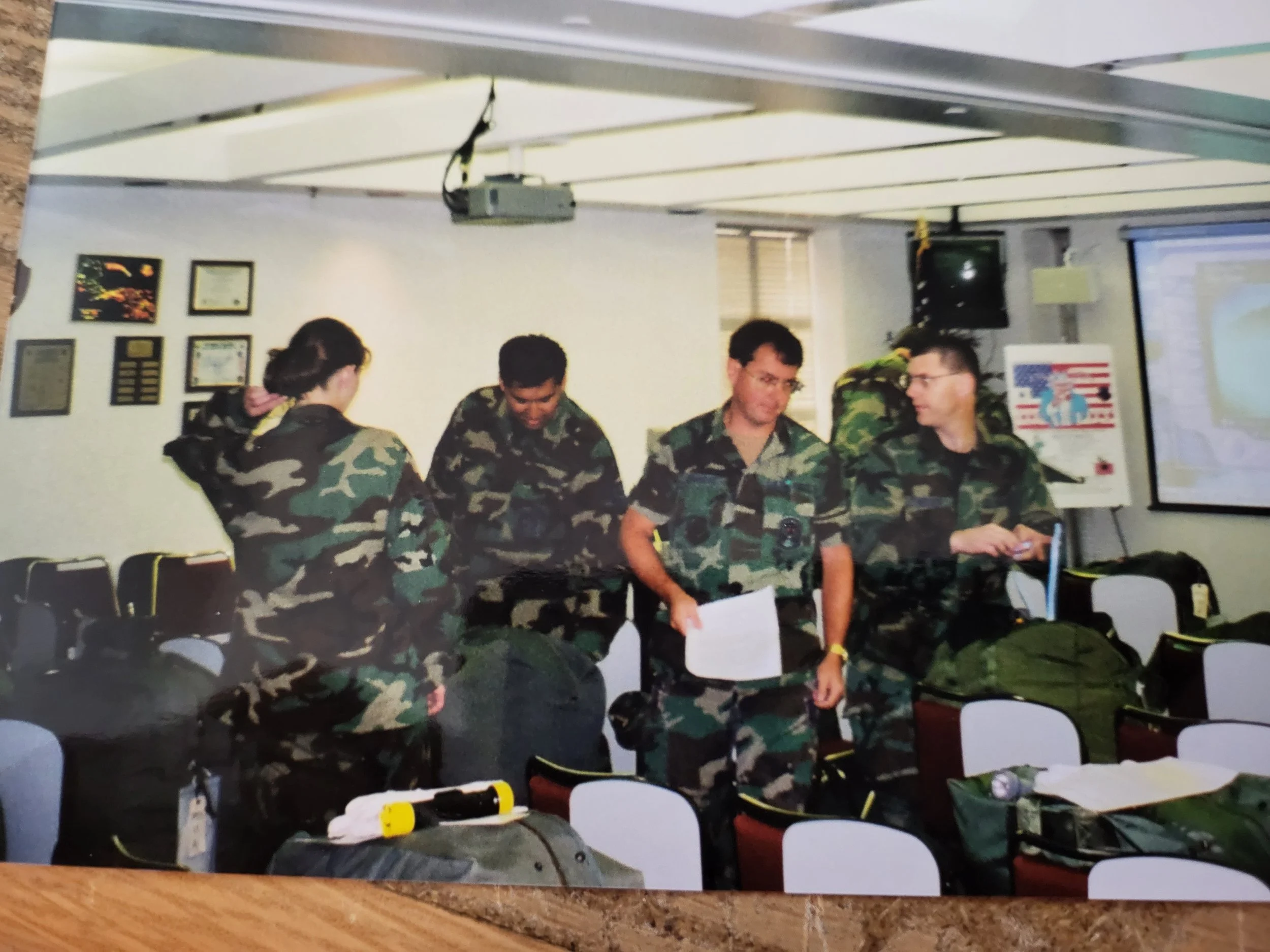 Military personnel in uniform standing in a classroom, some holding papers, surrounded by backpacks and equipment, with a presentation screen on the wall.