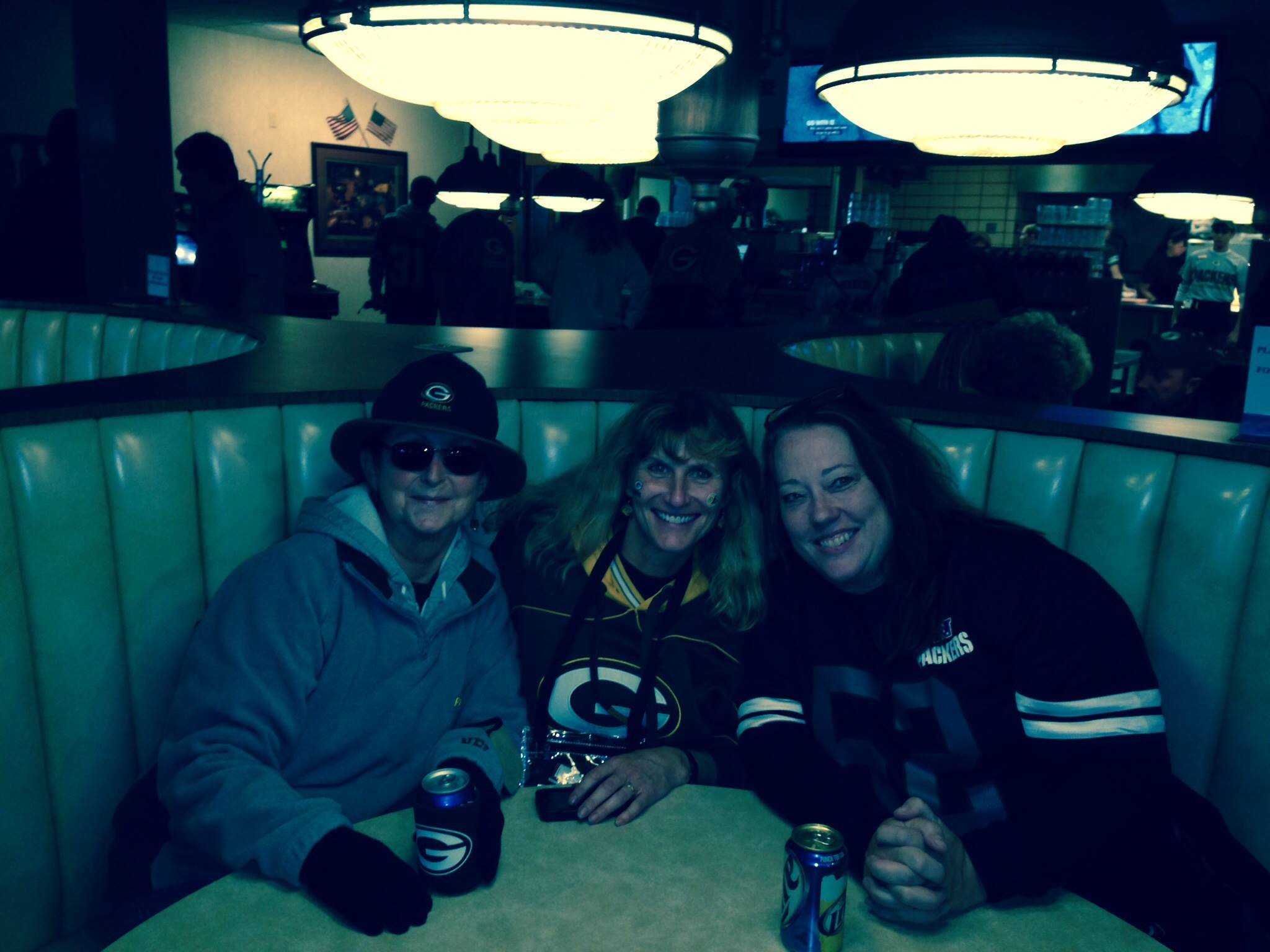 Three women sitting at a table in a restaurant, wearing Green Bay Packers apparel and holding cans of soda, with a background of dim lighting and other patrons.