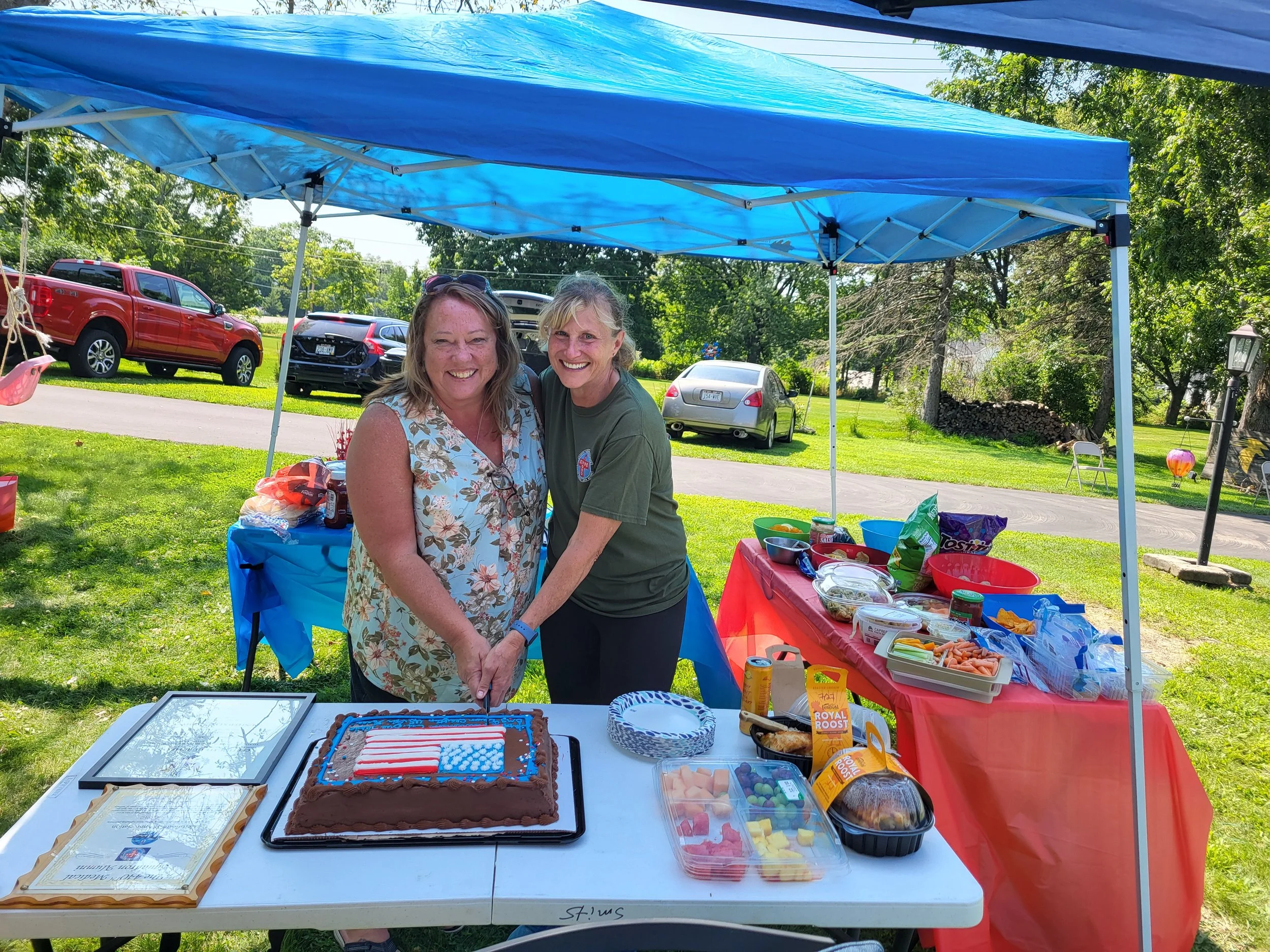 Two women smiling and standing behind a table with a firework-themed cake at an outdoor celebration, under a blue canopy with parked cars and green trees in the background.