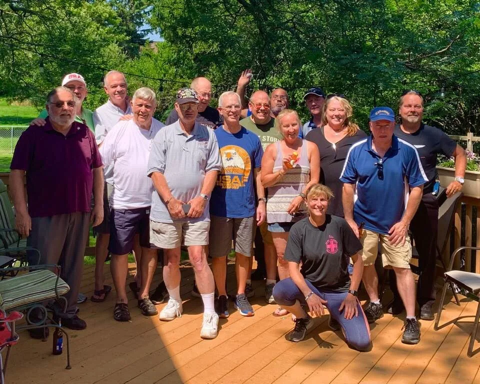 A group of fifteen people standing and kneeling on a wooden deck outdoors, surrounded by green trees, enjoying a sunny day.