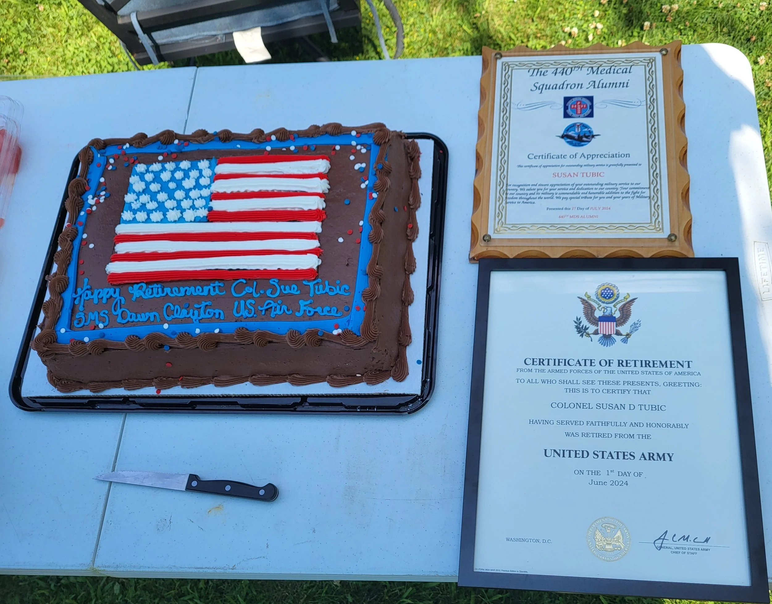 A celebration table with a chocolate cake decorated with an American flag design and a blue message that reads, 'Happy Retirement Col. Sue Tubic 3MS Ow 1st Lt. US Air Force.' There are two framed certificates of appreciation and retirement from the U