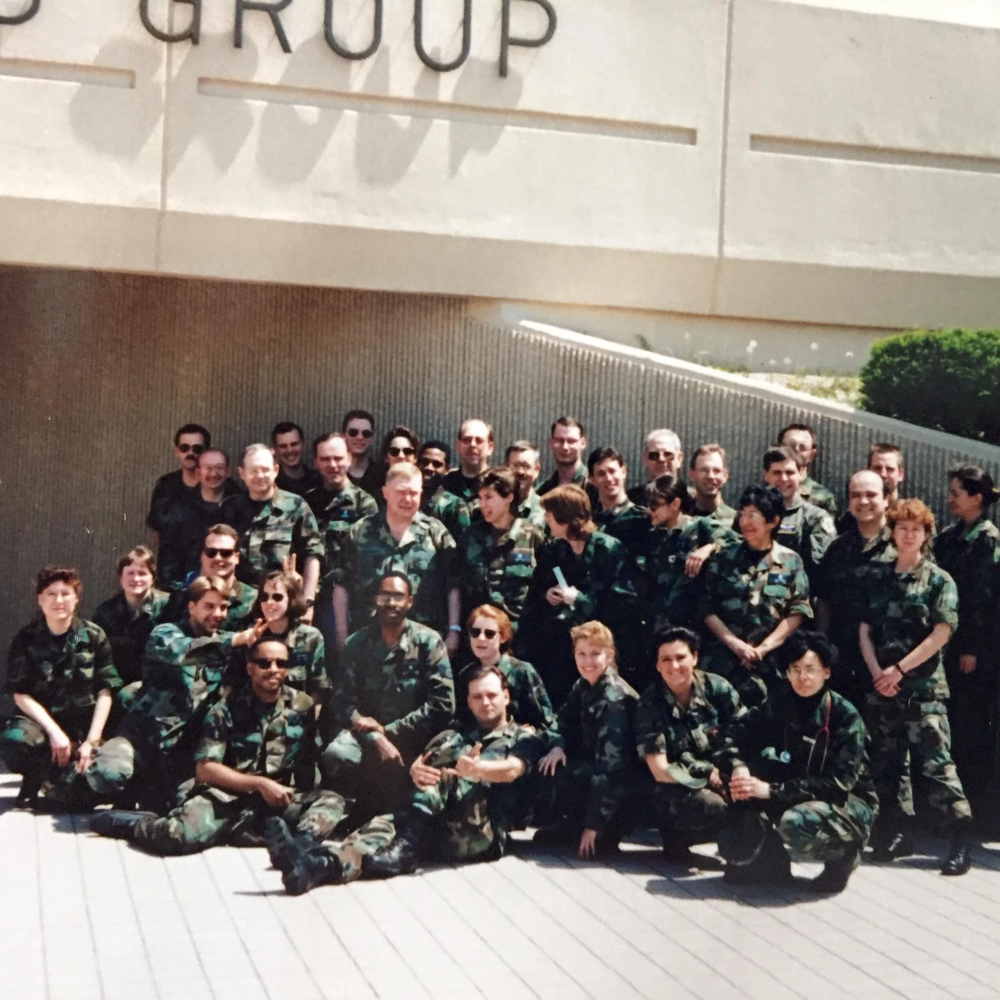 Group of military personnel in camouflage uniforms posing outdoors in front of a building with the word 'GROUP' on it.