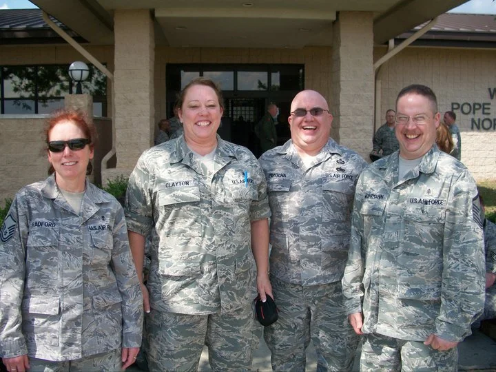 Four military personnel in camouflage uniforms standing outside a building, smiling.