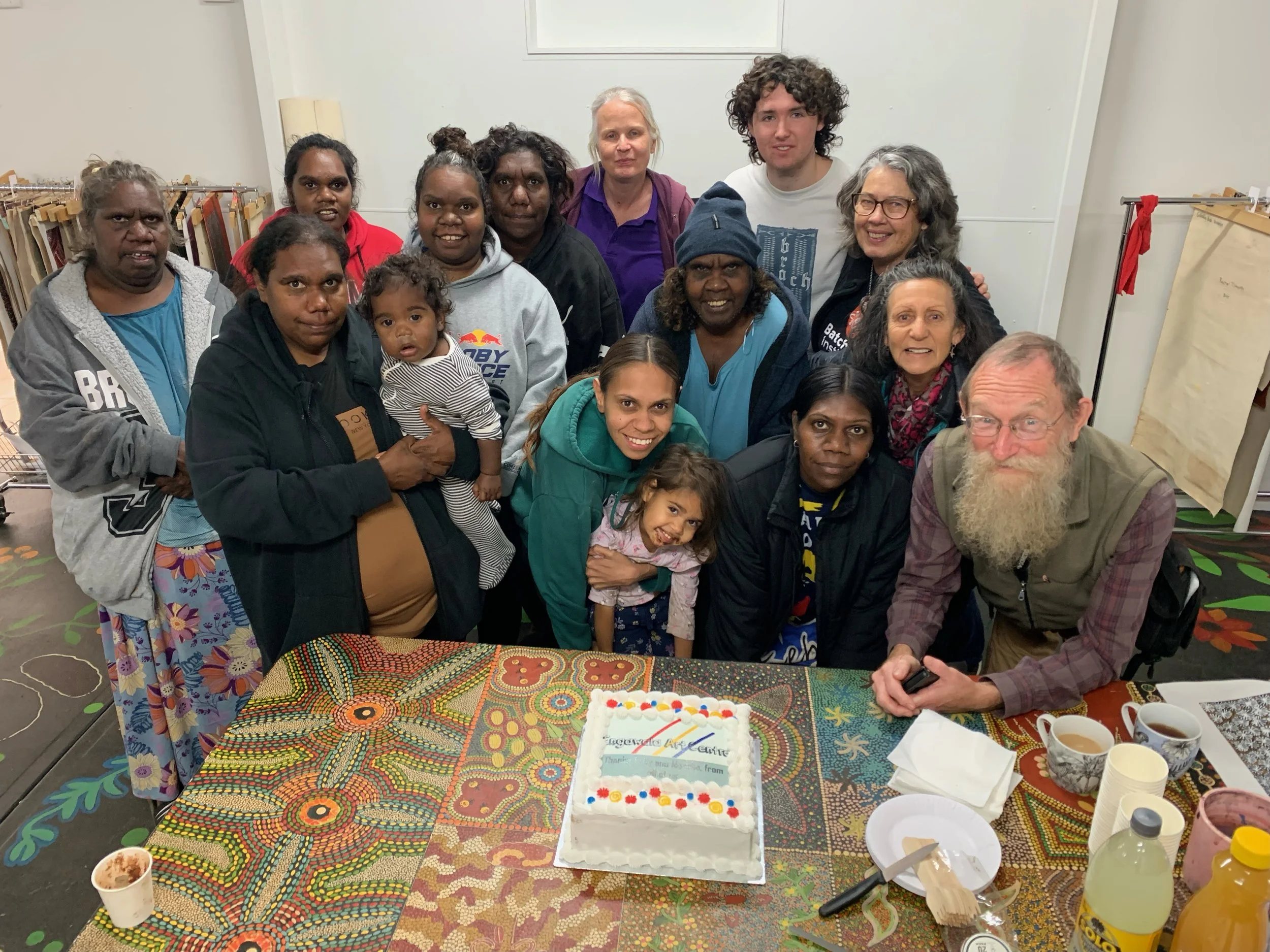 Group of diverse people celebrating around a table with a birthday cake, cups, and snacks in a community room.
