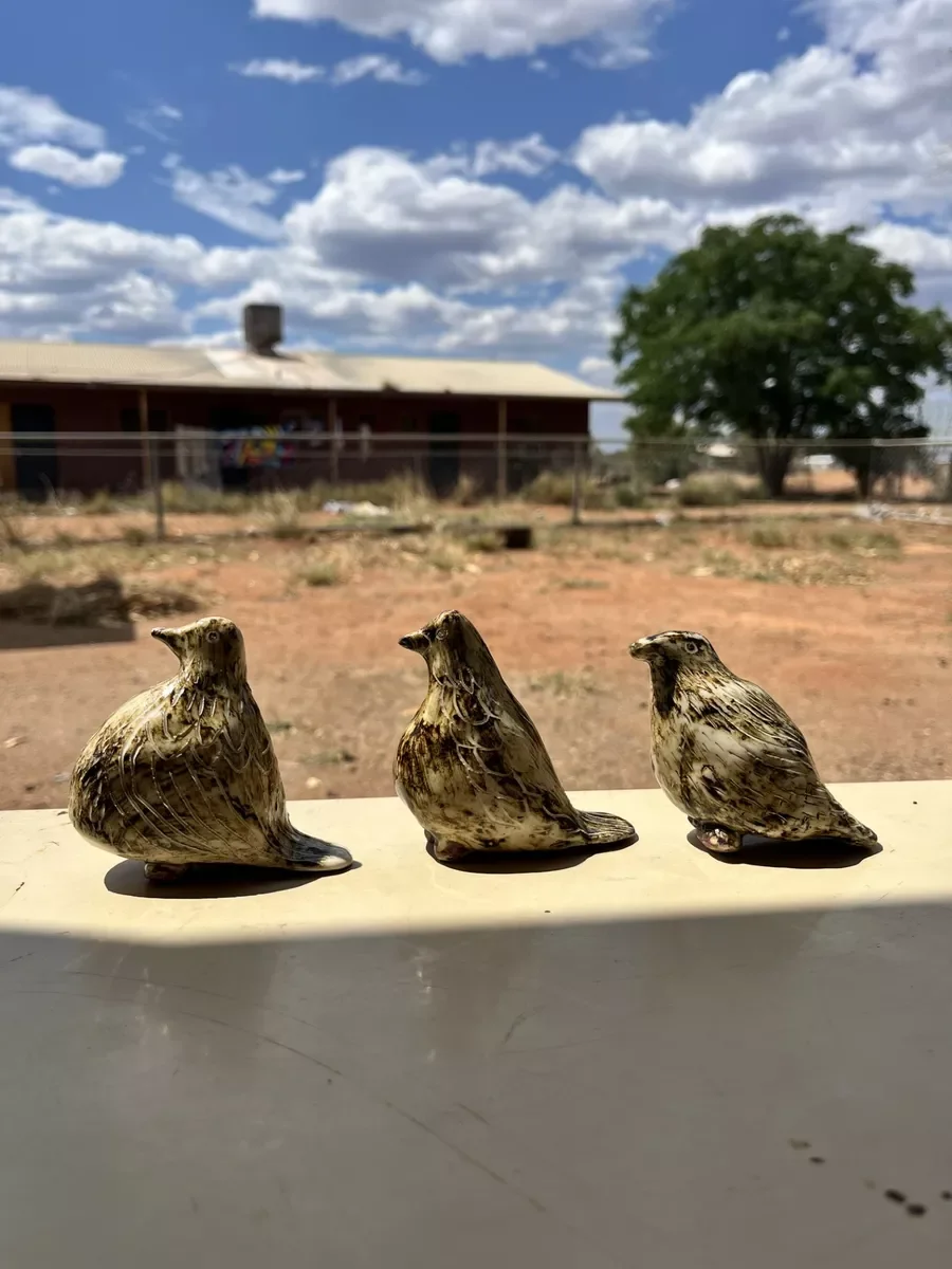 Three ceramic bird sculptures placed on a white surface outside with a house, a tree, and a cloudy sky in the background.