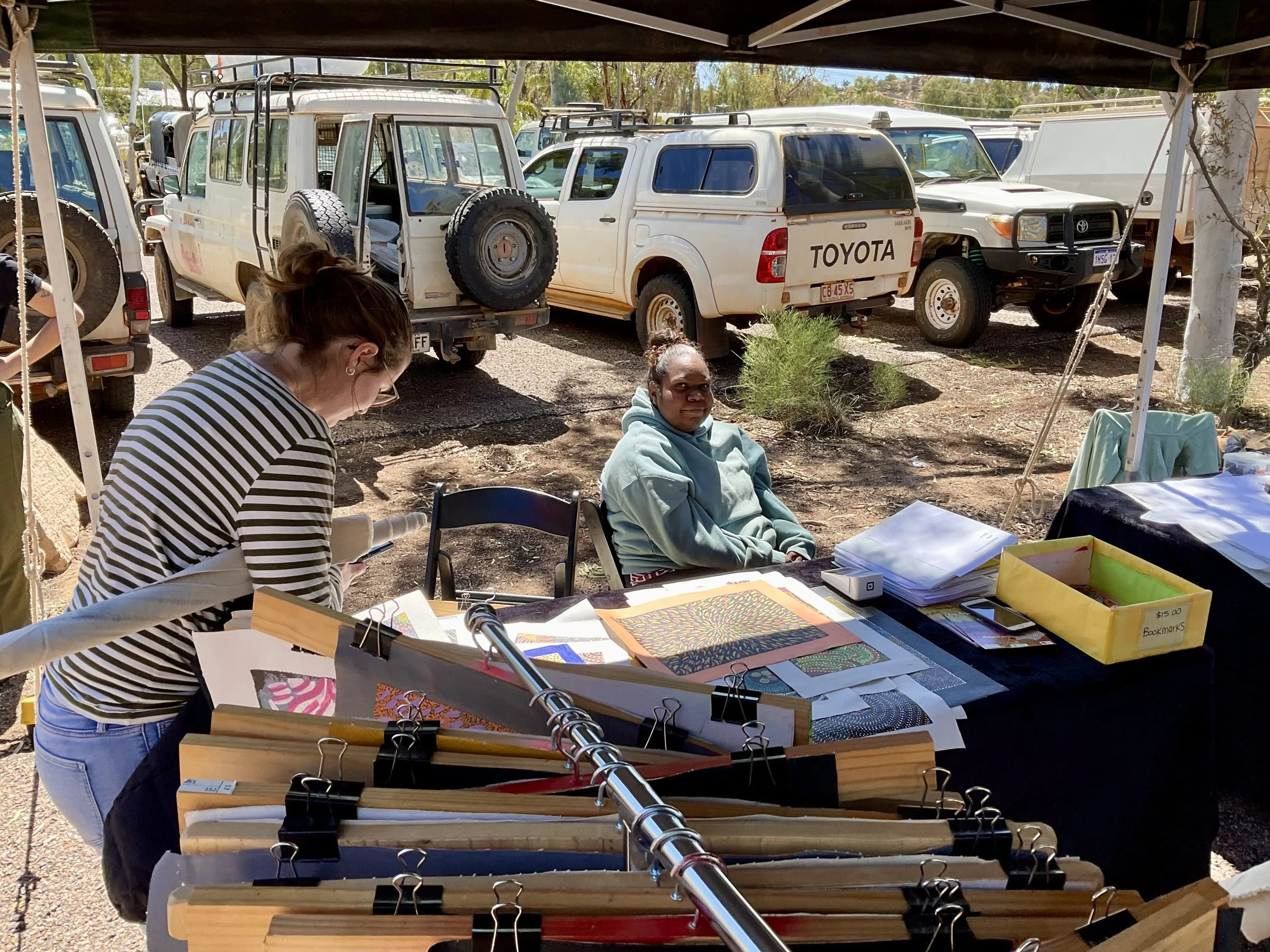 A woman in a striped shirt looking at papers on a table at an outdoor market or fair, with a woman sitting behind the table and several trucks parked behind her.