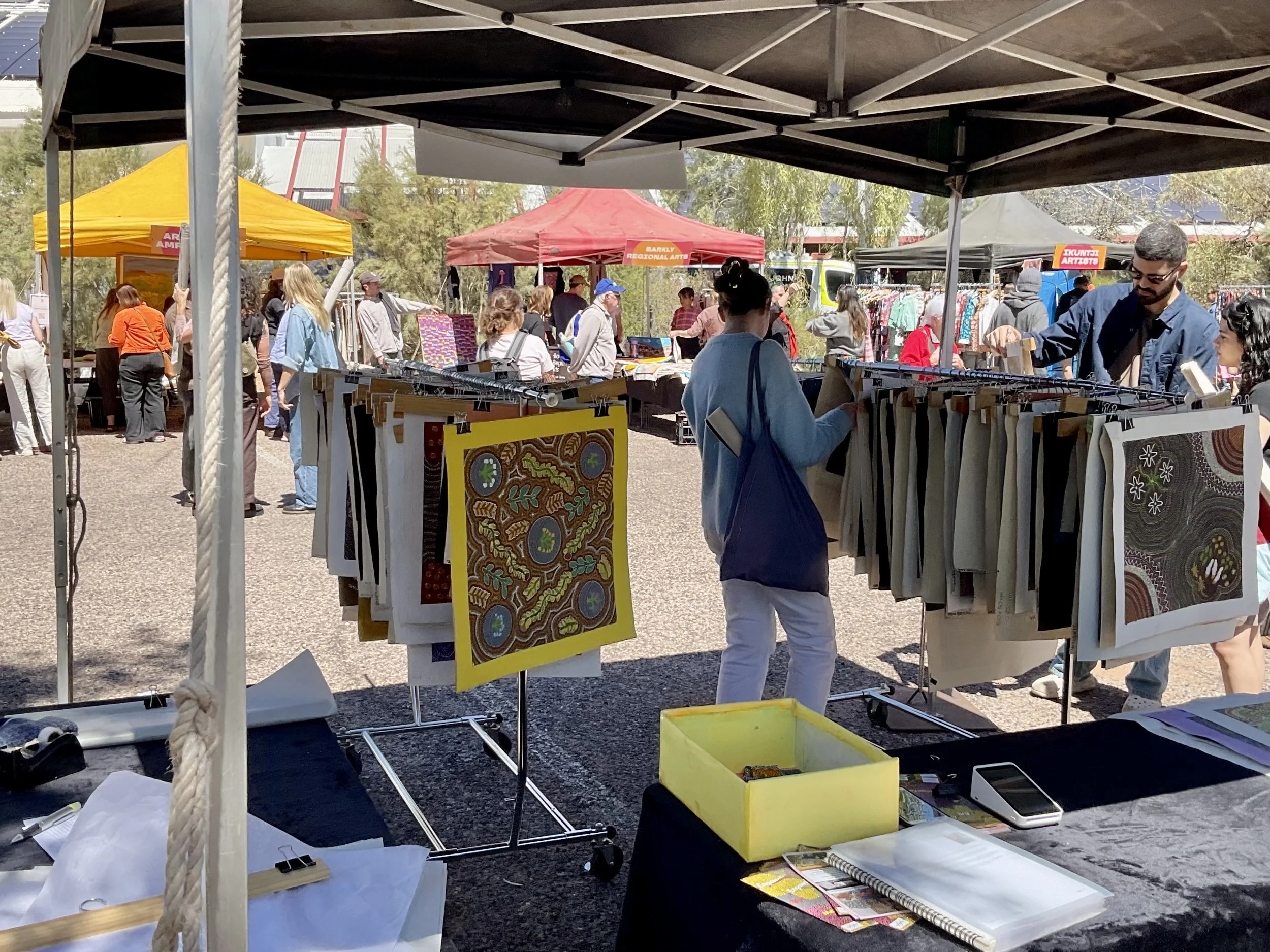 View of an outdoor arts and crafts market with people browsing various booths, including a stand selling paintings or fabric art. Several colorful tents and trees are visible in the background.