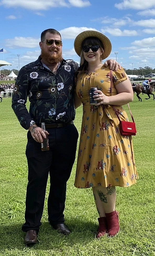 Image of Hayden and Cathy the owners of Smoke It Mackay at a Horse Race Day. Hayden wears a black and floral shirt with black pants. Cathy wears a mustard coloured floral dress