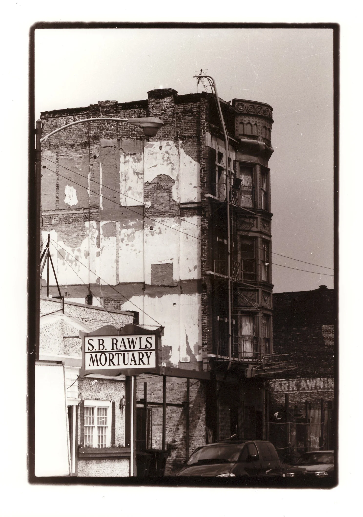 An old building with a brick facade and a rounded bay window at the top, with a sign in front reading "S.B. Rawls Mortuary." The image is in black and white.