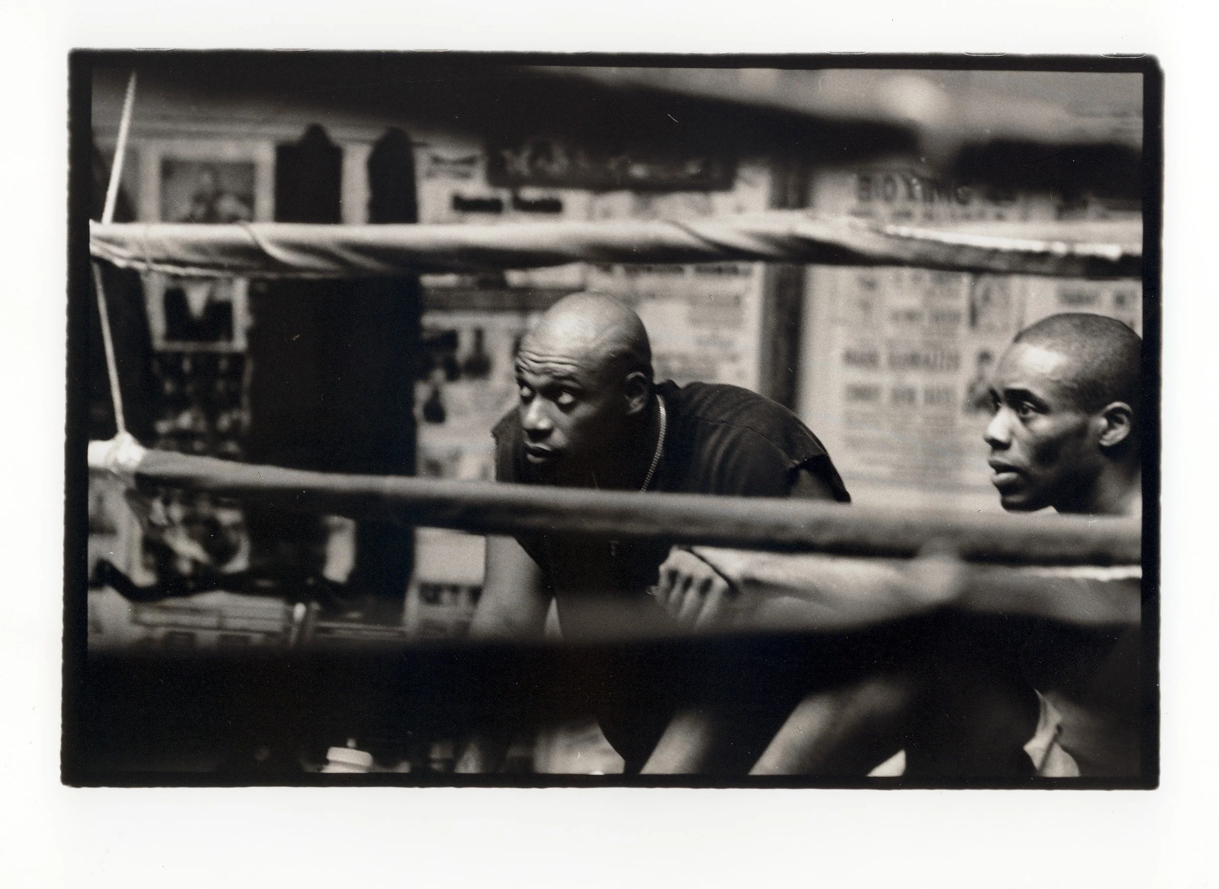 Black and white photo of two young men with shaved heads inside a boxing ring, with ropes in the foreground and a brick wall with signs in the background.