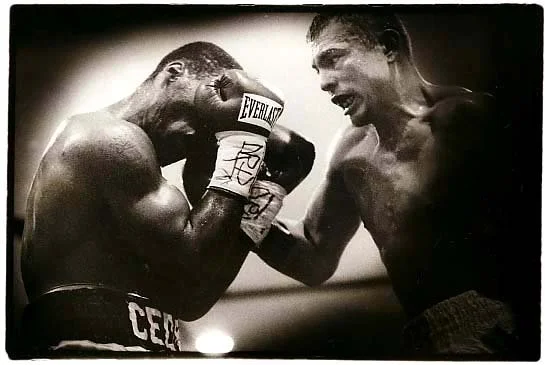 Two shirtless male boxers in a ring, engaging in a boxing match with one covering his face with gloves and the other smiling.