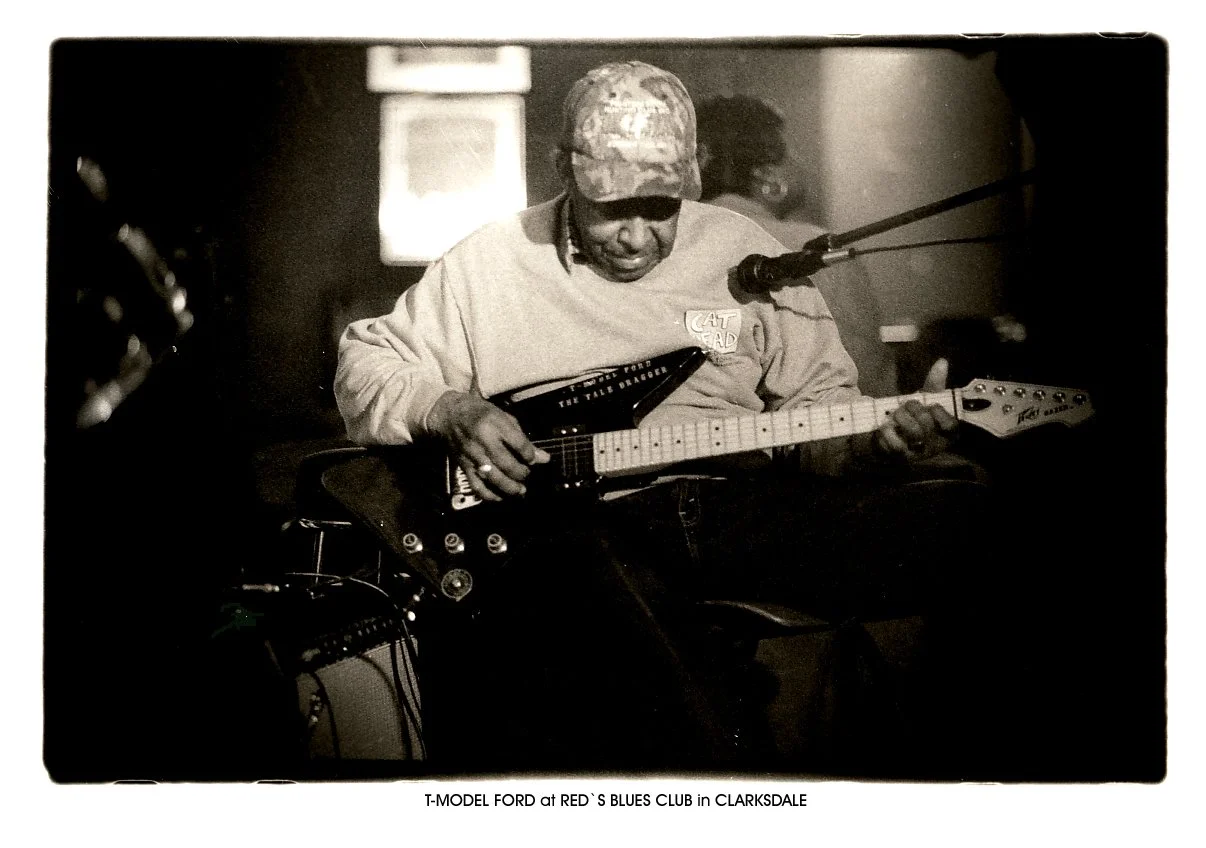 A man wearing a camouflage cap and a light-colored shirt, playing an electric guitar at a blues club in Clarksdale.