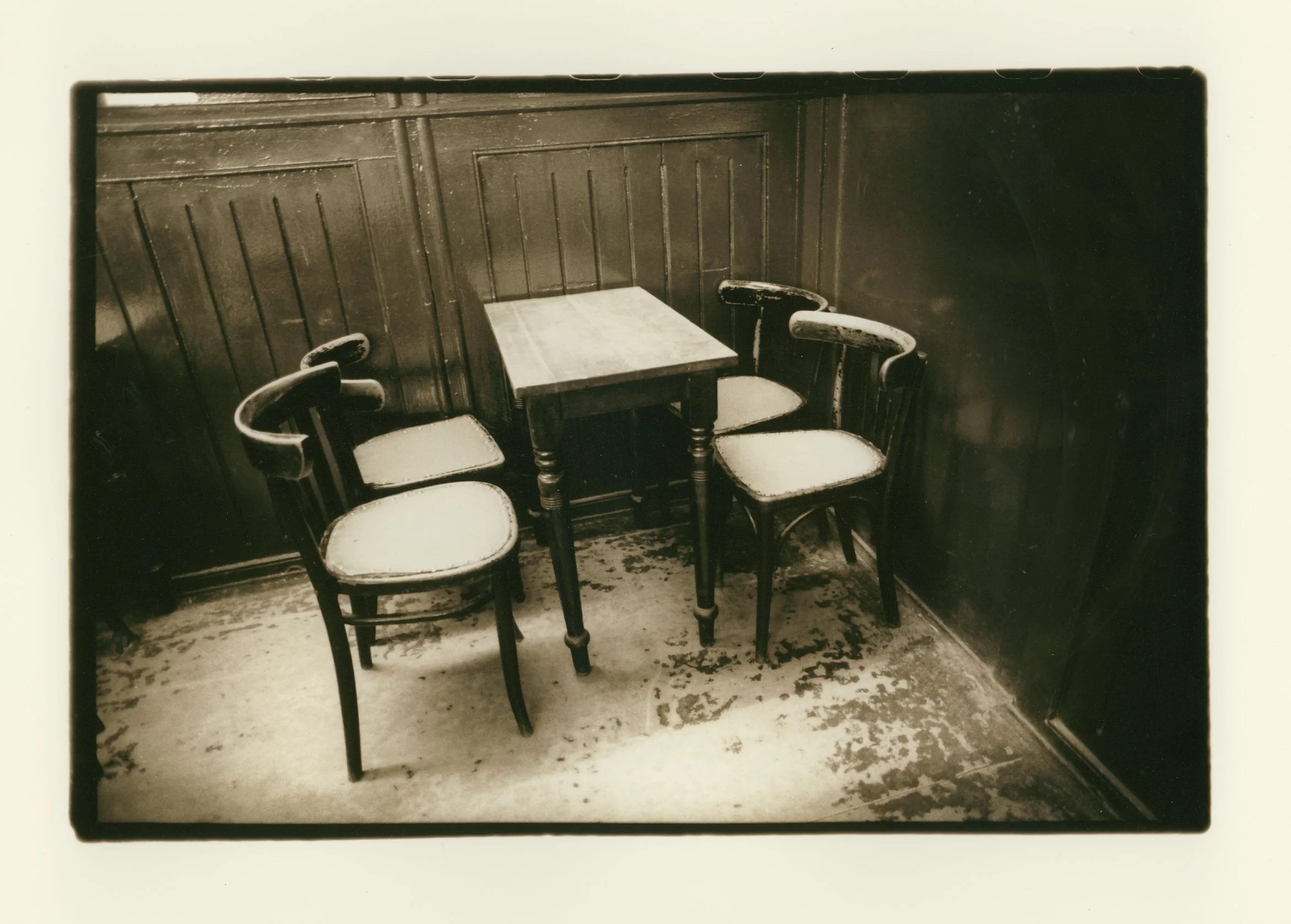 A vintage photo of a small wooden table with three chairs, set against dark wooden paneled walls and a worn floor.