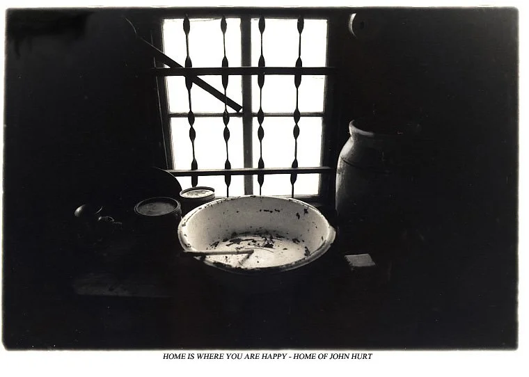 A rustic kitchen windowsill with a white dish, two jars, and a large ceramic pot silhouetted against a barred window, with a quote at the bottom that reads, 'Home is where you are happy -- home of John Hurt.'
