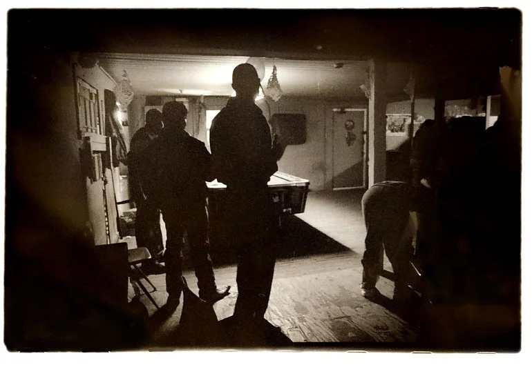 Silhouettes of people in a dimly lit room with a pool table in the background, some standing and others crouching.