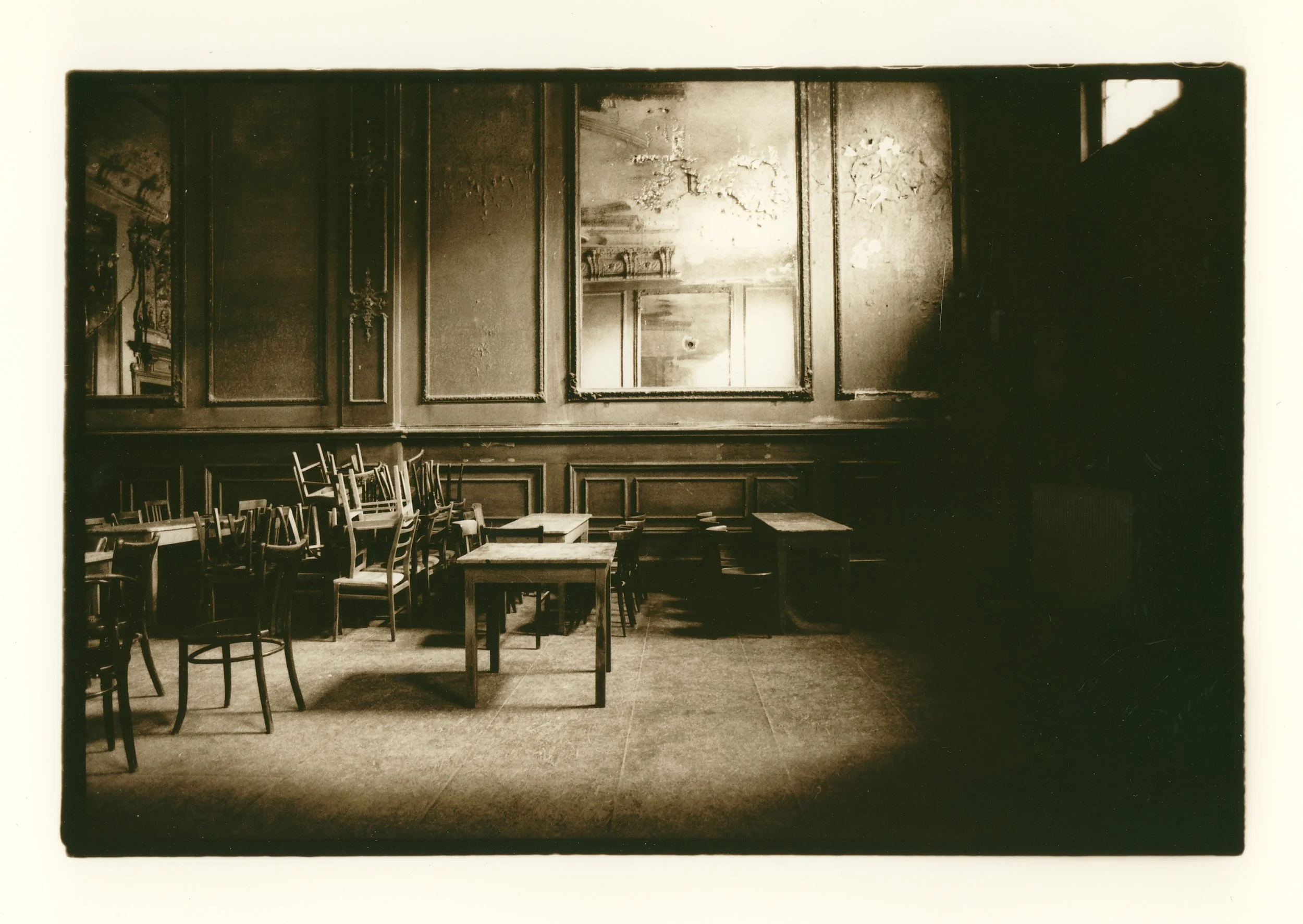 An old, abandoned room with worn wooden chairs stacked on top of each other and small tables. The walls have peeling paint and a large mirror or window is centered on the wall, reflecting parts of the room. The overall mood is dark and neglected.