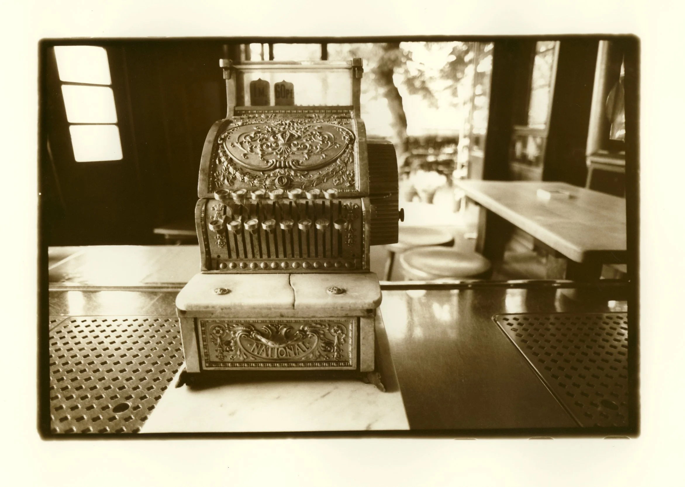 A vintage cash register with intricate decorative details on a wooden table inside a rustic setting, with windows and outdoor scenery visible in the background.